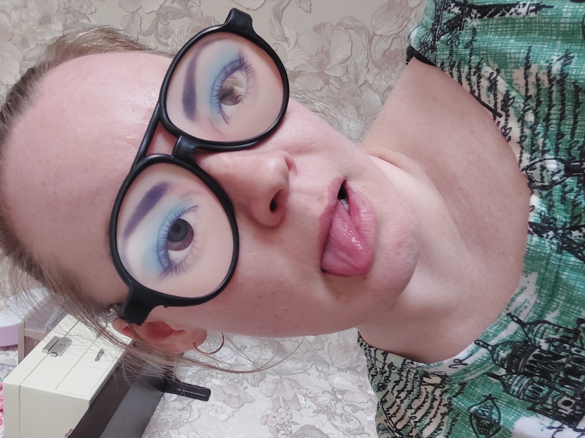 The image shows an adult person, likely female, captured in a close-up, rotated portrait. The individual is wearing large, black-rimmed, round glasses, through which their eyes, adorned with bright blue eyeshadow and long eyelashes, are visible. They are sticking their tongue out playfully, creating a mischievous expression. One ear is visible, featuring a simple gold hoop earring.
The person is wearing a green and white patterned shirt with black artistic renderings resembling buildings or architectural structures. The background consists of a light-colored, patterned wallpaper or fabric with ornate floral or damask motifs. To the lower left, a cream-colored box-like object with silver latches, possibly a jewelry box or small storage unit, is partially visible, next to a thin, dark rectangular object.
The scene appears to be indoors, likely a personal setting such as a home, given the wallpaper and personal items. The lighting is soft and even, suggesting either diffused natural light or typical indoor artificial lighting. No specific time of day or weather can be determined from the image. No discernible text is visible on any subject or object. The overall mood conveyed is lighthearted and informal. While the location context is Chaikovsky, Russia, there are no specific visual cues within the image itself to confirm this.