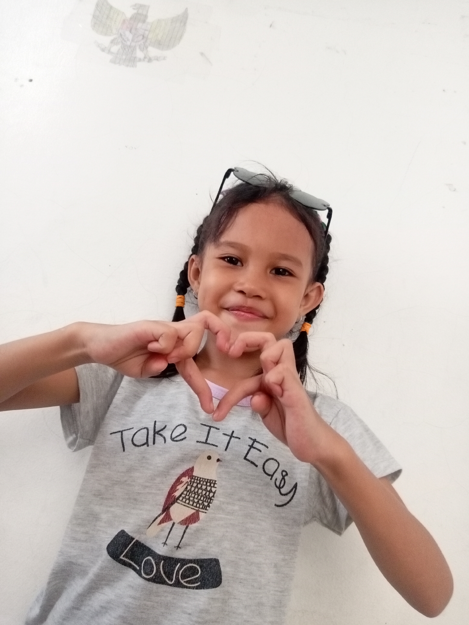 A young girl with dark hair in two braids, wearing a grey t-shirt with a bird graphic and the words "Take It Easy Love" printed on it, and sunglasses perched on her head, makes a heart shape with her hands in front of her chest. She is smiling and looking at the camera. The background is a plain white wall with a drawing of an eagle in the upper left corner. The location is a residential area in Batam, Indonesia.