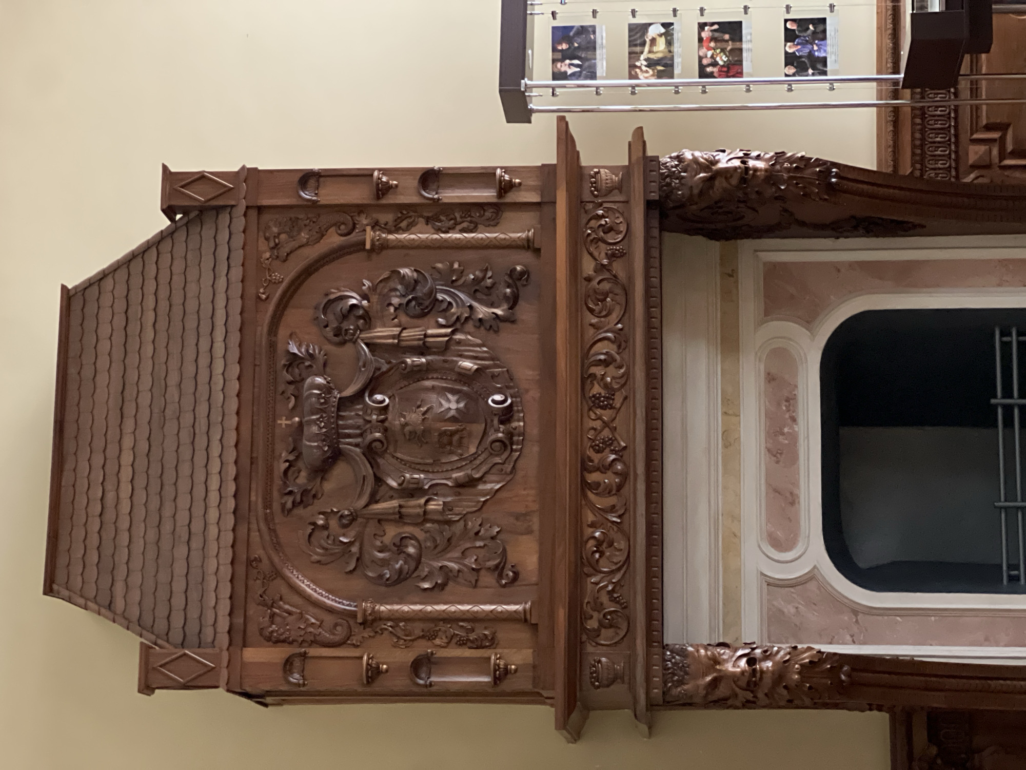 The image shows an elaborately carved wooden fireplace in Saint Petersburg, Russia. The fireplace features a large, ornate crest with intricate scrolling and a crown at the top. The mantelpiece is also decorated with detailed carvings, including floral motifs and faces on the supports. Above the crest, the fireplace extends upwards into a structure resembling a roof, also made of wood. To the right of the fireplace, a glass display case contains several framed photographs of people, possibly historical figures or performers. The background is a plain, light-colored wall. The fireplace appears to be a significant decorative element in what is likely a historical building or museum.
