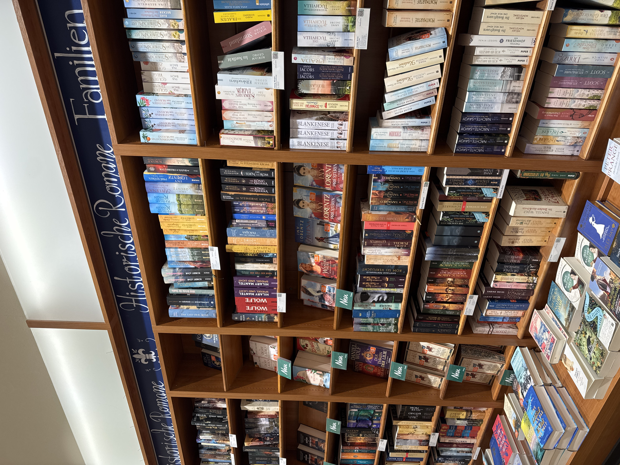 The image shows a bookstore interior, specifically the book section dedicated to "Historische Romane" (Historical Novels) and "Familien" (Family) books. The shelves are made of warm-toned wood and are fully stocked with a variety of books. The books are arranged in rows on multiple shelves, with some standing upright and others stacked horizontally. Many of the books have colorful covers, while others have more subdued designs. Several books are displayed open, showcasing their pages. Small green "Neu" (New) tags are visible on some of the shelves, indicating new arrivals. The overall environment appears clean and well-organized, typical of a retail bookstore. The lighting is bright, suggesting an indoor setting. The text "Historische Romane Familien" is written in white cursive script on a dark blue banner above the shelves. This bookstore is located in the city of Münster, Germany.