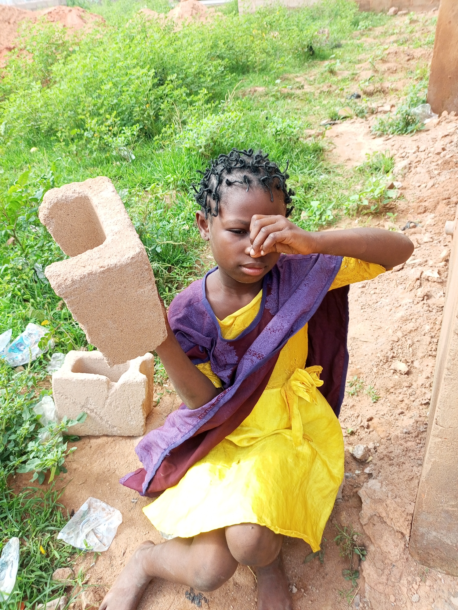A young girl, with dark skin and braided hair, sits on the dirt ground outdoors. She wears a yellow dress and a purple and maroon wrap. Her right hand is raised, covering her eyes, and she appears to be rubbing them. In her left hand, she holds a tan-colored cinder block. In front of her, on the ground, is another similar cinder block, partially submerged in the dirt. The background is filled with green foliage and a brown, dusty surface. Scattered around the girl are pieces of white plastic trash.