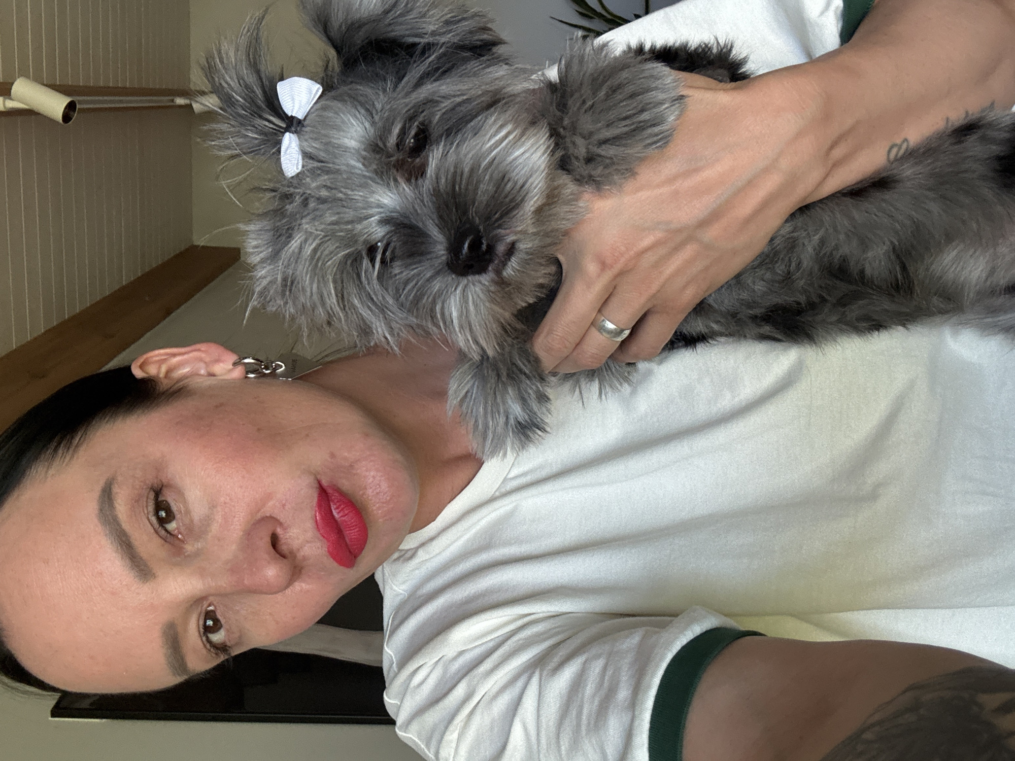 A woman is holding a small, grey dog with a white bow in its hair. The woman is wearing a white t-shirt with green trim and has dark hair pulled back. She is wearing red lipstick and earrings. The dog is held close to her face. The location is Chaikovsky, Russia. The environment appears to be indoors.