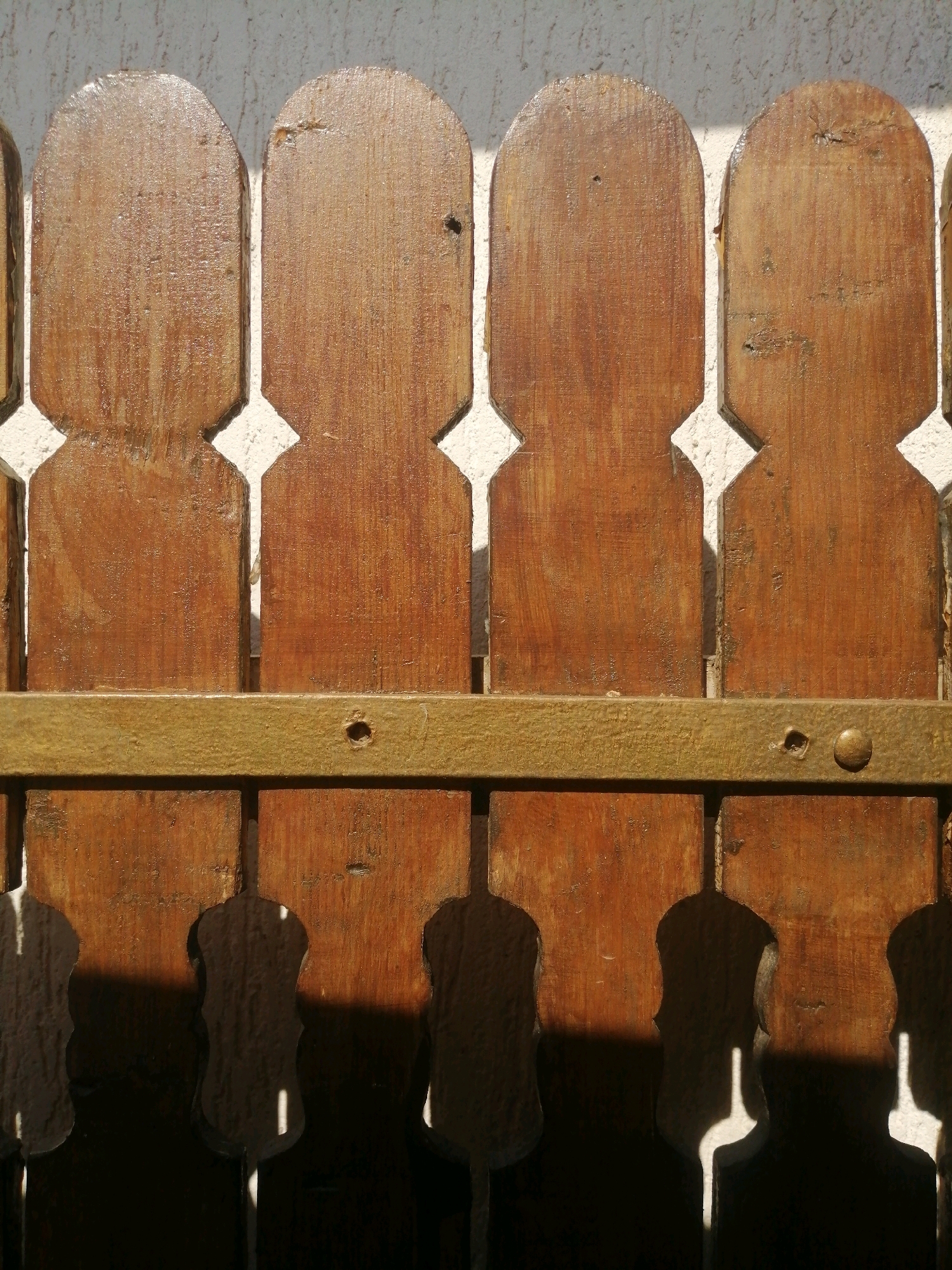 The image shows a close-up, eye-level, outdoor shot of a wooden fence with decorative cutouts. The fence is made of vertical wooden planks with rounded tops, and the lower portion of each plank has an hourglass-shaped cutout. A horizontal metal bar runs across the middle of the fence, fastened with rivets. The background is a textured white wall. Sunlight casts shadows through the cutouts, creating a pattern on the ground below, which is not fully visible. The wood appears weathered and stained. The scene is in an unknown location in an unknown city.