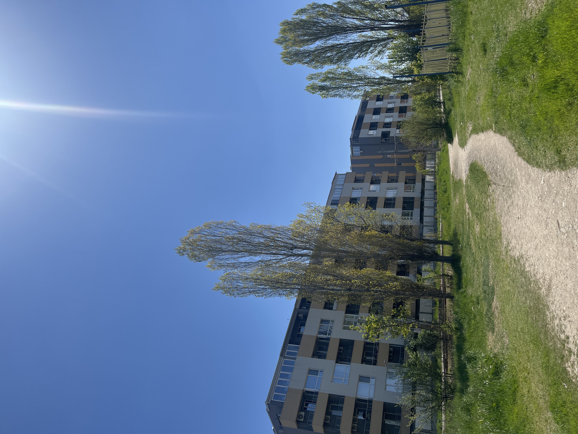 The image shows a street scene in Sofia, Bulgaria, with a gravel path running alongside a grassy area and a modern apartment building. The sky is clear blue with bright sunlight. Trees with sparse spring foliage line the path. A metal fence is visible between the trees and the path in the background. The apartment building has a striped facade with a mix of light-colored and dark-colored panels and windows. There are no people visible in the image. The overall impression is of a sunny day in a residential area.
