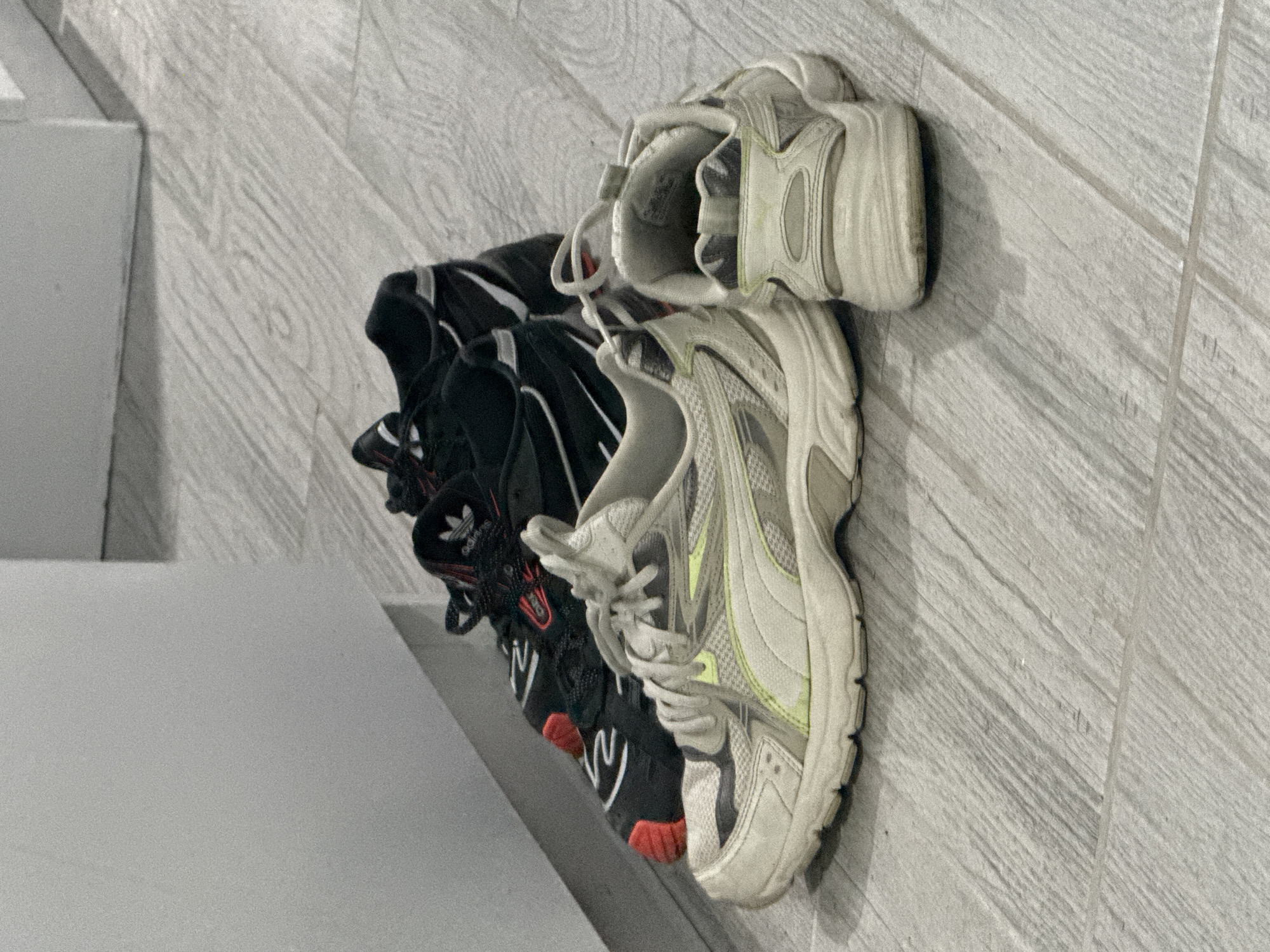 The image shows three pairs of sneakers stacked on a grey, tiled floor. The bottom pair is black with red accents and the Adidas logo visible. The middle pair is white and grey with lime green details. The top pair is also white and grey. The image does not contain information to infer its location as Stara Zagora, Bulgaria.
