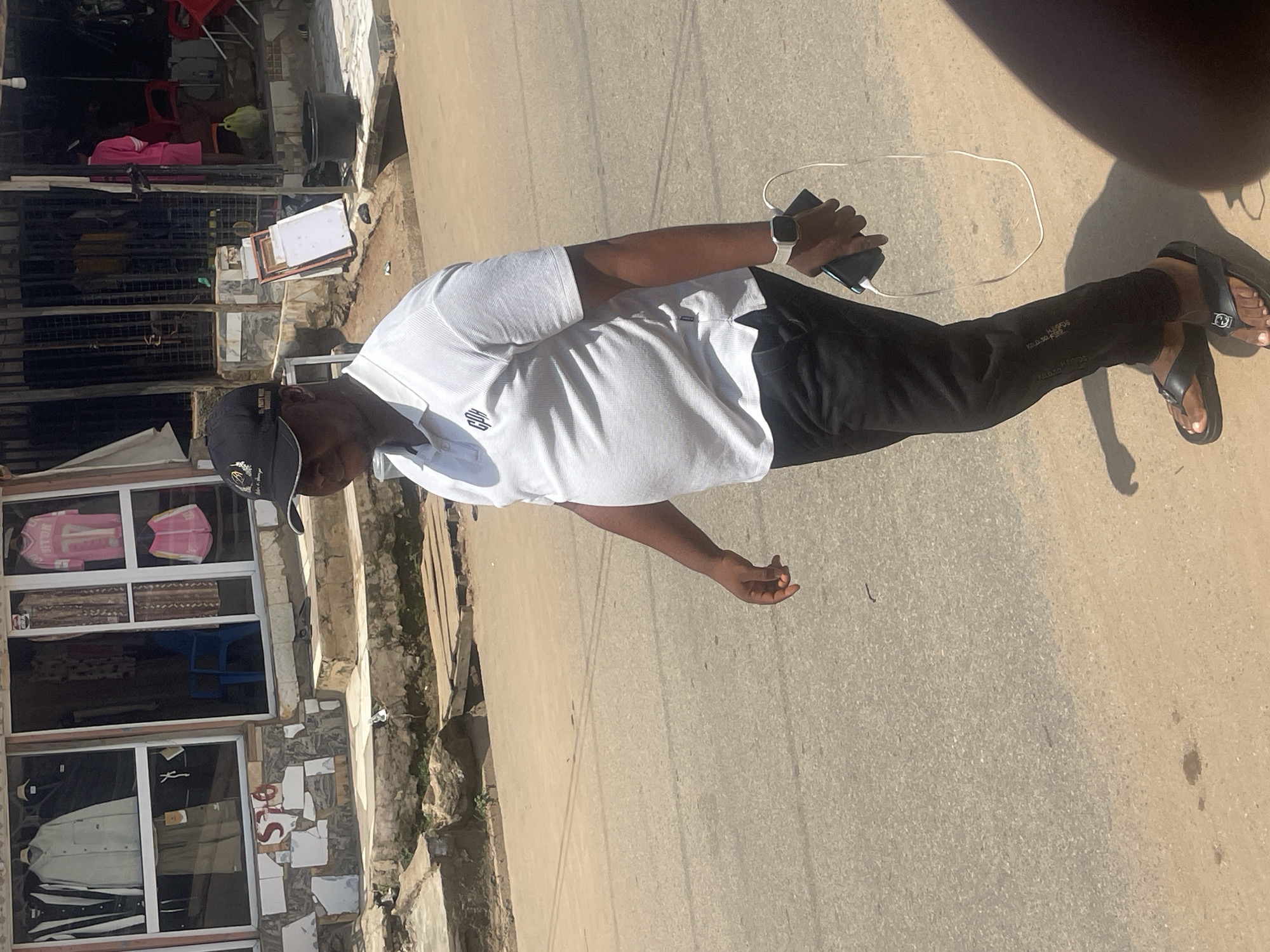In Abuja, Nigeria, a man of dark complexion wearing a white polo shirt, black pants, and black flip-flops walks along a paved street in front of a store. He is holding a black smartphone in his right hand, which is connected by a white wire to a smartwatch on his left wrist. The store has a tiled facade with various items displayed in its windows, including clothing and what appear to be mannequins. A blue chair is visible inside the store. The street is bordered by a drainage ditch and a sidewalk. The overall scene is bathed in daylight, suggesting an outdoor urban environment.