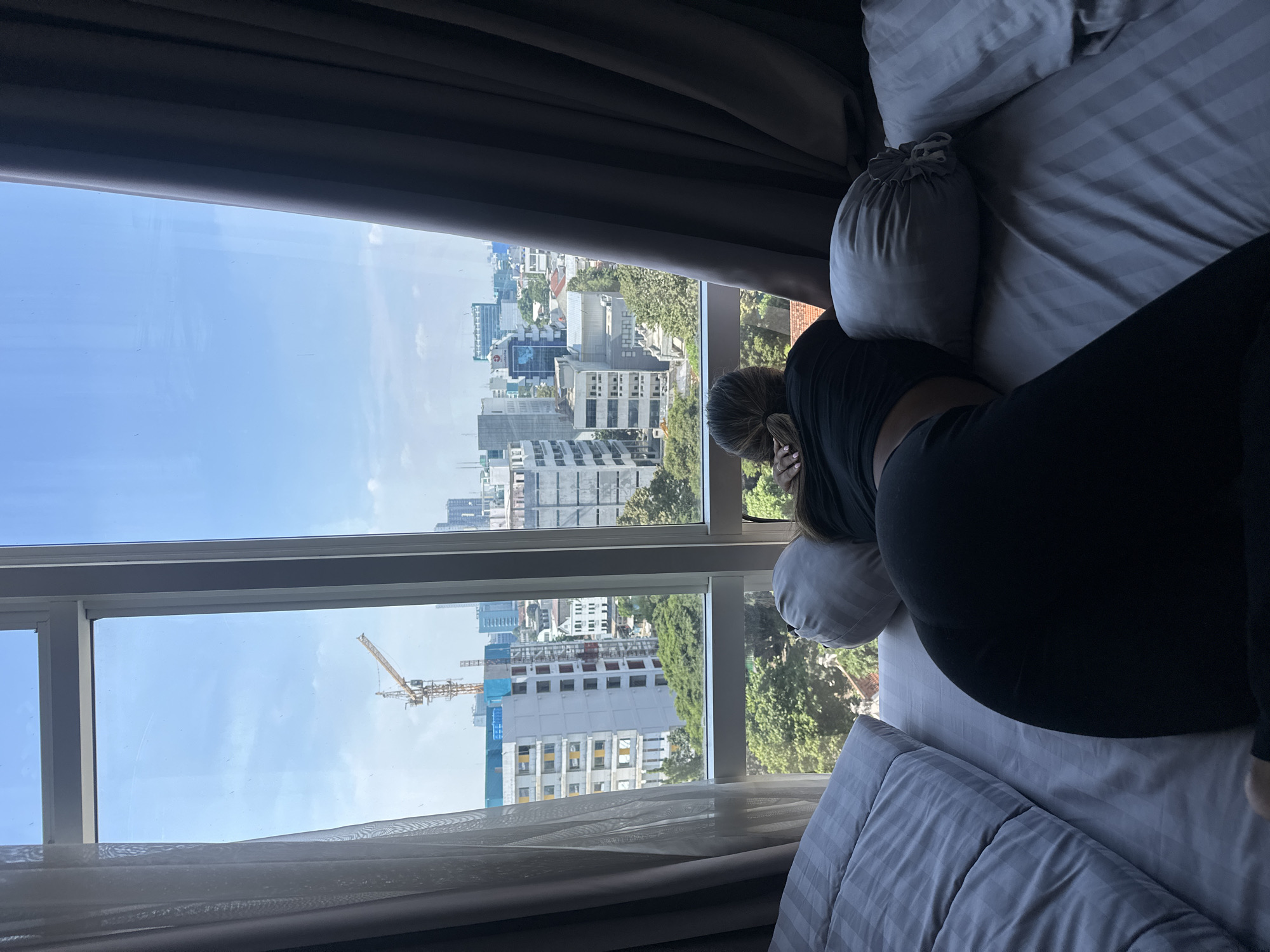 A woman lies face down on a gray striped bed near a window. The window offers a view of a city with buildings under construction, featuring a crane. The city can be identified as Central Jakarta, Indonesia based on the architecture and urban environment.