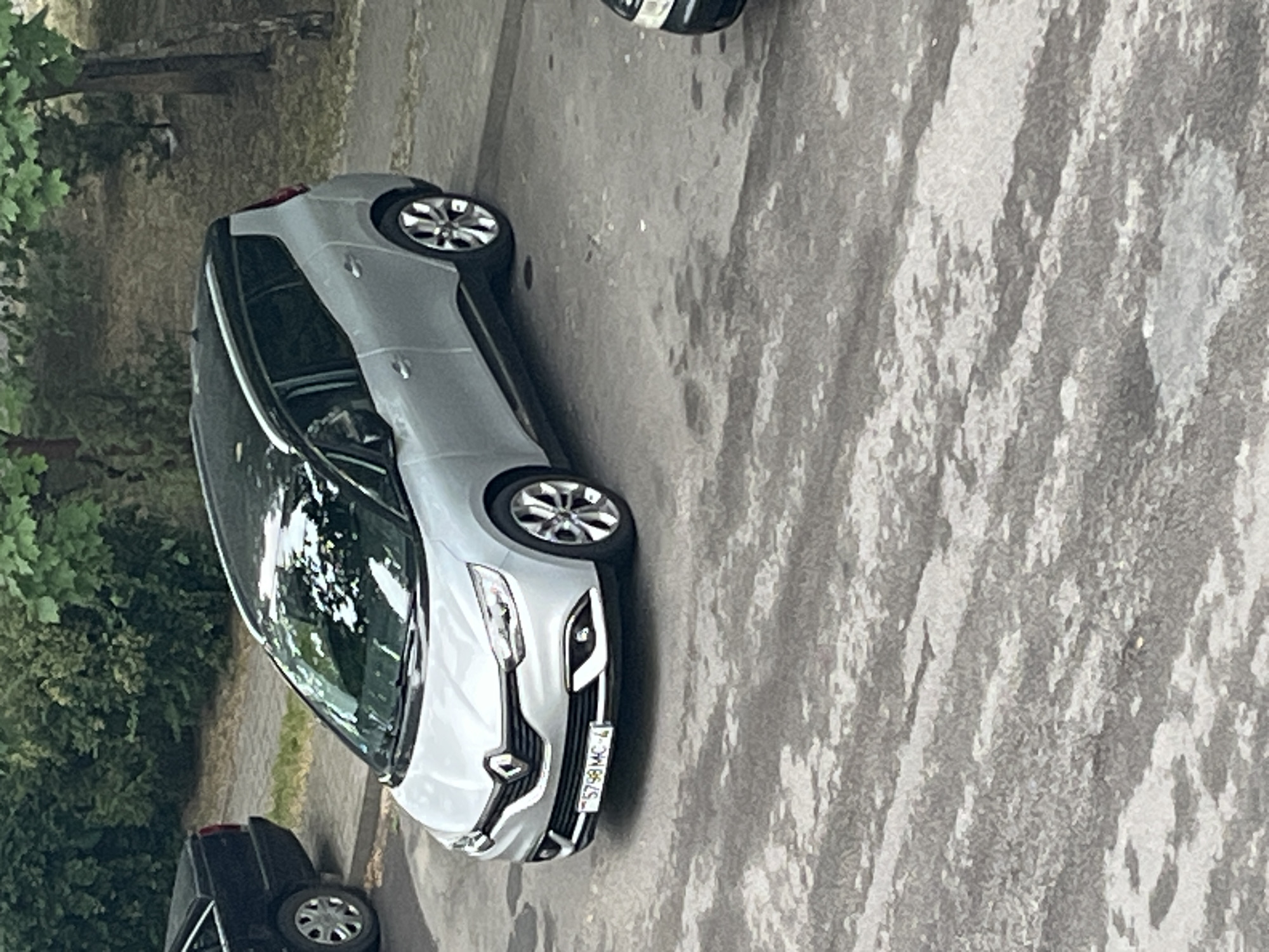 The image, rotated 90 degrees counter-clockwise, displays an outdoor scene featuring two cars on a paved surface, bordered by green foliage.
The primary subject is a light-colored (likely silver or light gray) car, prominently positioned in the foreground and center. It appears to be a Renault model, identified by the diamond-shaped logo on the front grille. The car has modern styling with sleek headlights and alloy wheels. Visible on its front license plate is the text "5798 MC-4".
In the background, to the lower left, a dark-colored car is partially visible, showing its rear wheel and a portion of its body. The ground beneath the vehicles is a paved surface, possibly asphalt or concrete, with varying shades and textures, suggesting wear or different materials.
To the left and in the deeper background, there are numerous green trees and bushes, indicating a natural or landscaped environment surrounding the paved area. The lighting suggests it is daytime, with no strong shadows to indicate direct sunlight, possibly an overcast day or the light filtered through trees. There are no people visible, and no specific event or activity is taking place other than the cars being parked or stationary.