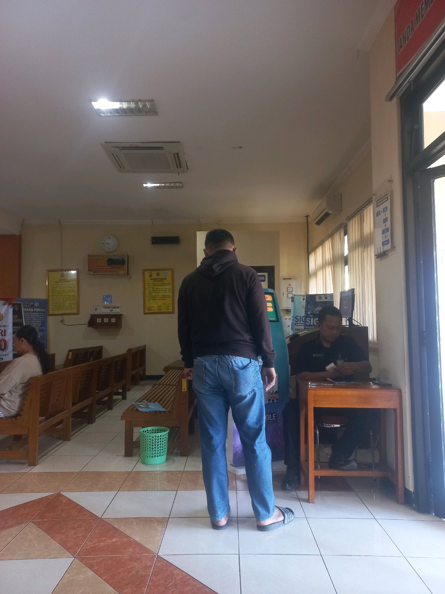 The image depicts the interior of an office or service area in Baturetno, Indonesia. In the foreground, a person wearing a black hooded sweatshirt and blue jeans stands with their back to the camera, facing a counter area. To the left, another person is seated on a wooden bench, facing away from the camera. Several rows of wooden benches are visible, suggesting a waiting area. In the background, a man in a dark uniform sits behind a wooden desk, appearing to be attending to paperwork. A computer monitor and a sign with text are also visible on the counter. The room has a tiled floor and a drop ceiling with fluorescent lighting and an air conditioning unit. A red sign with white text is partially visible at the top right of the frame, and a window is present to the right. Several framed posters or notices are displayed on the walls.
