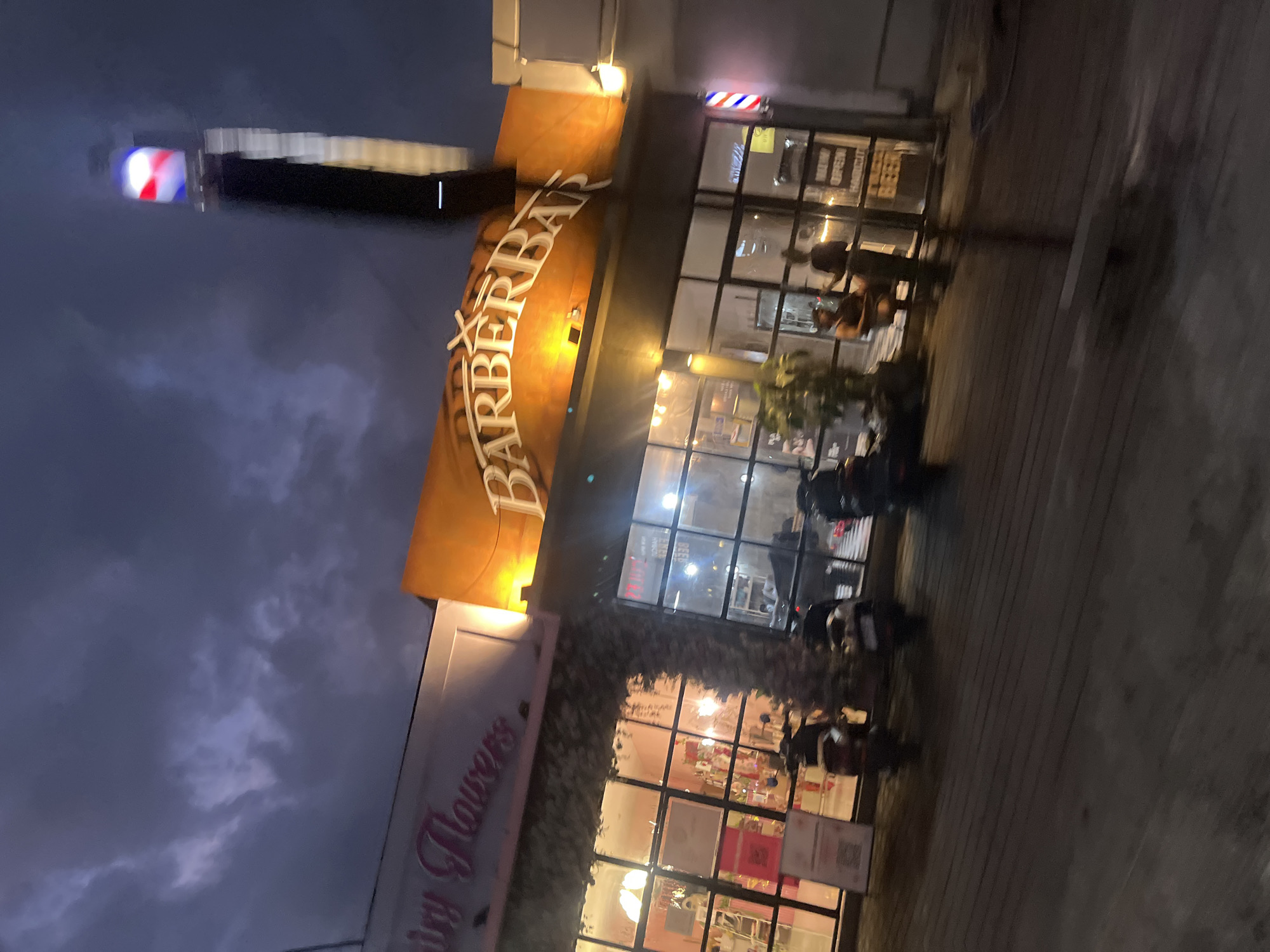 This image depicts a street scene at dusk in North Kuta, Indonesia. The primary focus is on the exterior of two storefronts. The first, prominently featuring a large, orange awning with the white text "BARBERBAY" in stylized script, suggests a barbershop. Above the awning, a classic red, white, and blue barber pole is visible, further confirming its purpose. The building has large glass windows, allowing a glimpse of the interior, which appears to be well-lit.
Adjacent to the barbershop is another establishment, seemingly a flower shop, with a sign that reads "Sunny Flowers" in pink script. This storefront also has extensive glass windows, revealing a bright, pink-toned interior with floral arrangements or products displayed inside.
Several parked motorcycles are visible outside the shops, indicating a common mode of transportation in the area. A few people can be seen near the entrances, possibly customers or staff. The sky is a deep, dusky blue with visible clouds, suggesting the transition from day to night. The lighting from the storefronts casts a warm glow on the pavement. The overall atmosphere is that of a commercial street during the evening hours.