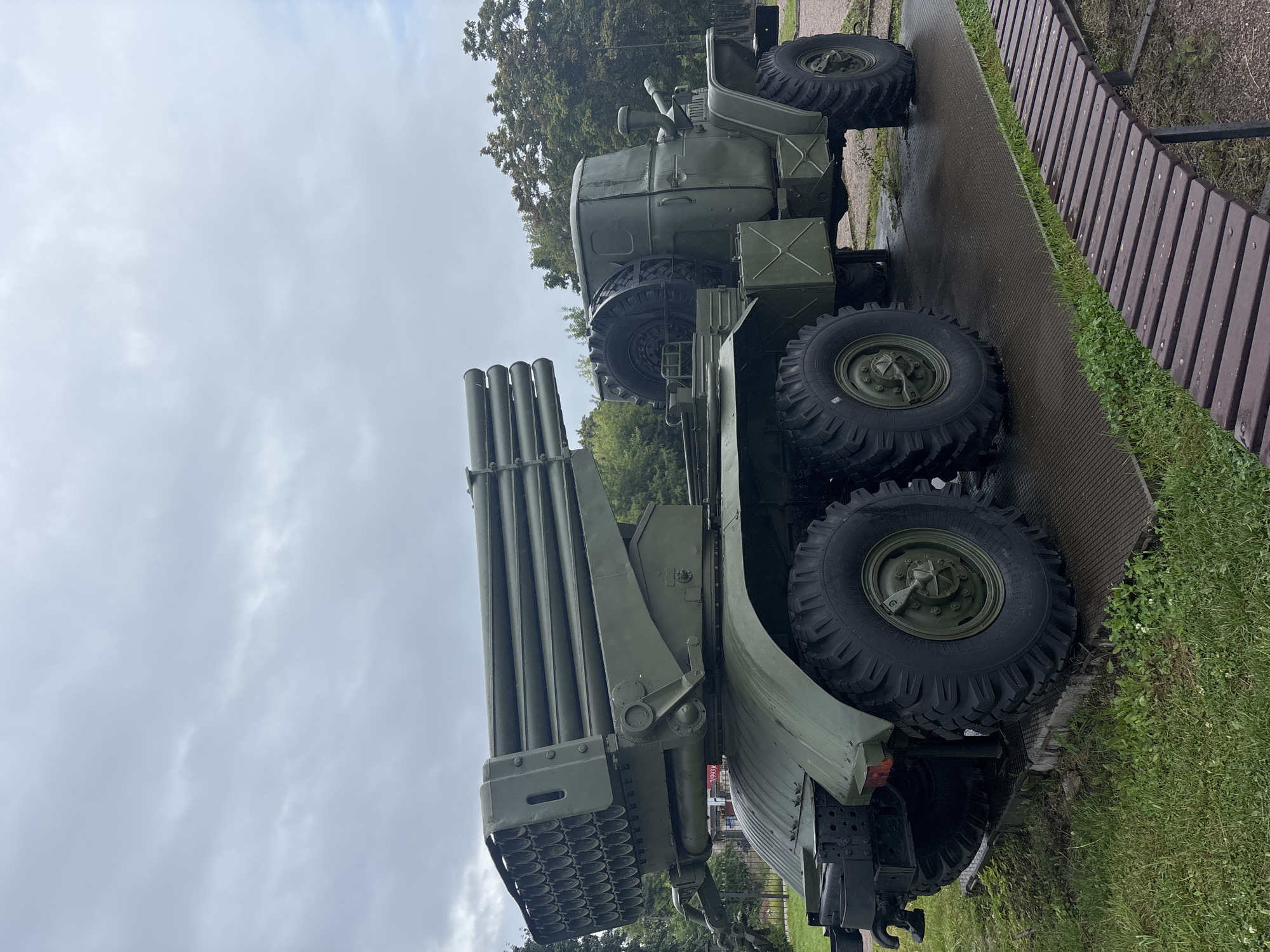 The image shows a Soviet-era BM-21 Grad multiple rocket launcher, painted in olive drab, displayed outdoors on a cloudy day. The vehicle is positioned on a paved area next to a grassy patch and a wooden bench. The launcher, with its characteristic multiple tubes, is prominently featured at the rear of the truck. In the background, trees and a chain-link fence are visible. A sign partially visible behind the launcher appears to have Russian text. This artifact is likely located at a memorial or museum related to military history in the city of Luga, Russia. There are no people or animals present in the image. The environment appears to be an outdoor exhibition space.