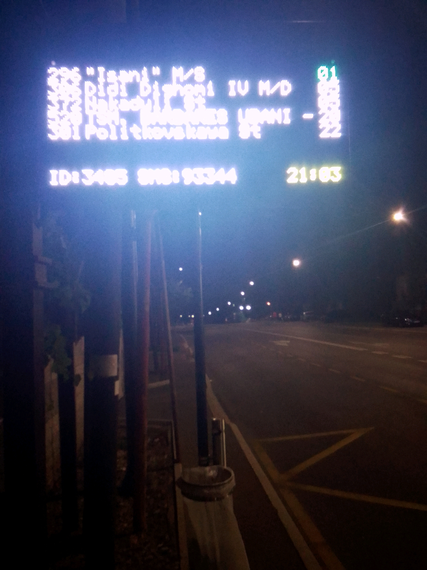 A digital bus stop display is visible at night in Tbilisi, Georgia. The display shows a route number "296" followed by " "Isani" M/S Didi Dighomi IU M/D". Below this, it lists "Natsarkeuli St" and "Usmani - 21". Further down, it reads "Sol Polikhovskaya St - 22". At the bottom left, "ID: 3435 SMS:93344" is displayed, and to the right, "21:03" is shown. The display is illuminated with bright white and green light, with some parts appearing slightly out of focus or overexposed. The background shows a dark street scene with streetlights and faint outlines of buildings and trees. A portion of the bus stop pole and a trash receptacle are visible in the foreground on the left side of the image. The ground appears to be asphalt with yellow road markings.