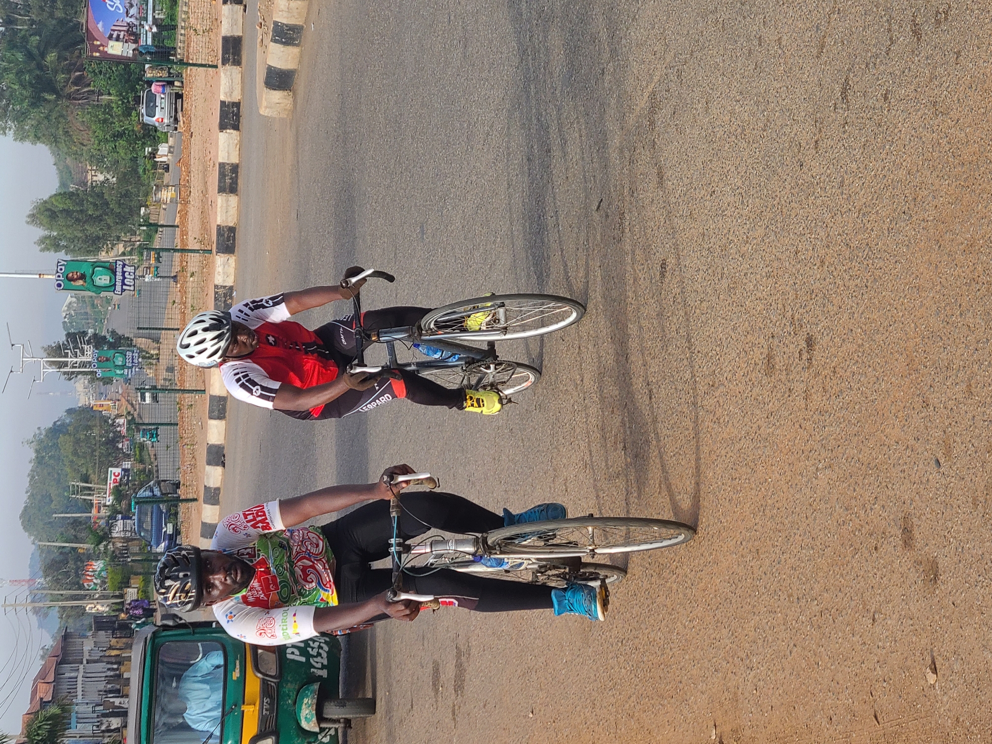 Two cyclists ride on a paved road in Gura Top, Nigeria. One wears a red and white jersey with black tights, a white helmet, and yellow shoes. The other wears a white jersey with colorful accents and black tights, a helmet, and blue shoes. Both are riding racing bicycles. A green auto rickshaw is visible to the left. In the background are buildings, signs, trees, and hills. The road is edged with black and white striped barriers.
