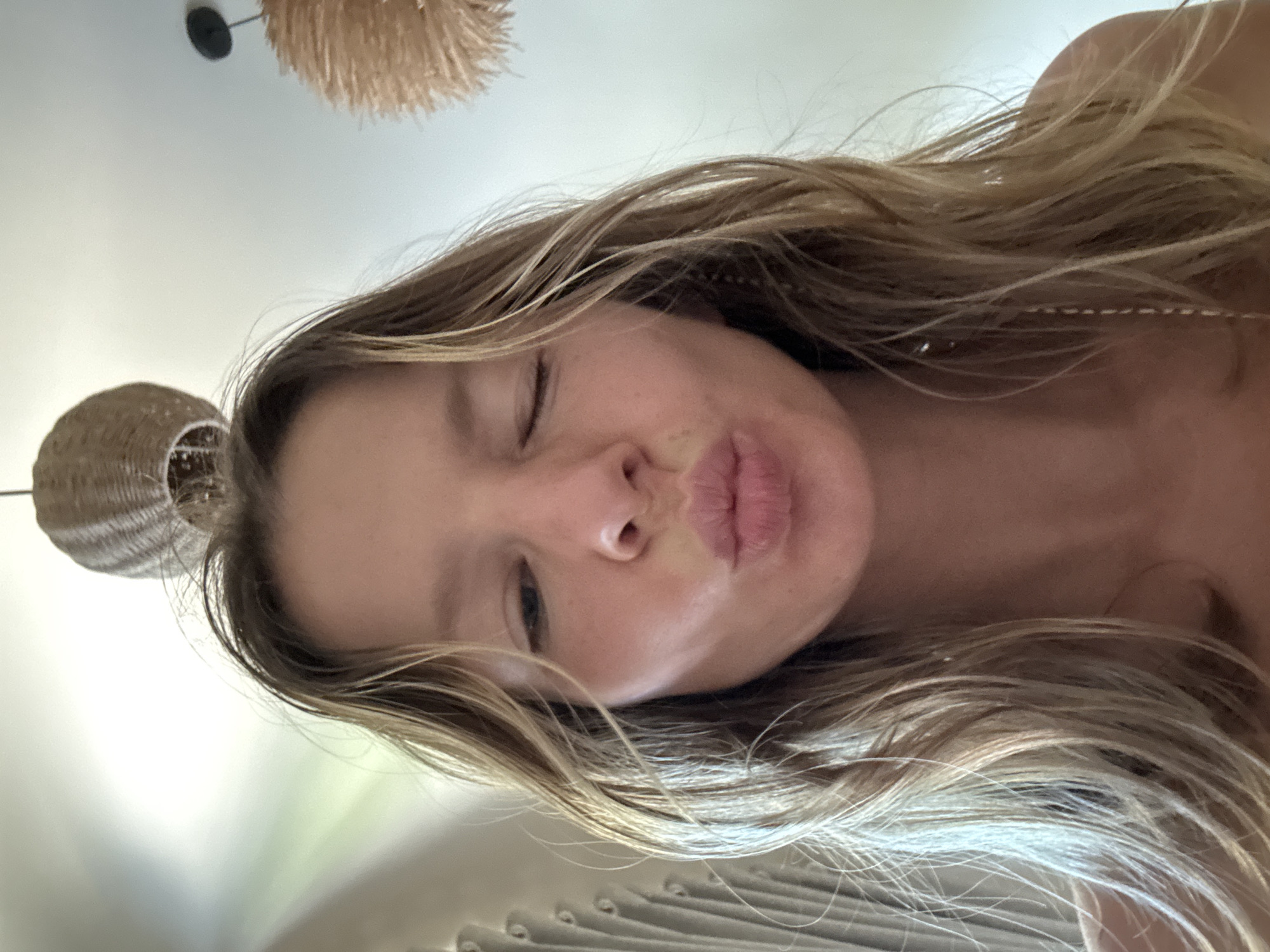 A medium close-up selfie shows a young woman with long, wavy blonde hair, winking with her left eye and puckering her lips. Her skin is fair with a smattering of freckles. The background is softly blurred, with a hint of a thatched lampshade above and a textured curtain to the left. The lighting is bright and natural, creating a warm glow. The scene suggests an indoor setting, possibly a bedroom or living area. The woman's pose and expression convey playfulness and a casual demeanor. There is no indication of a specific location in Mũi Né, Vietnam, or any urban environment.