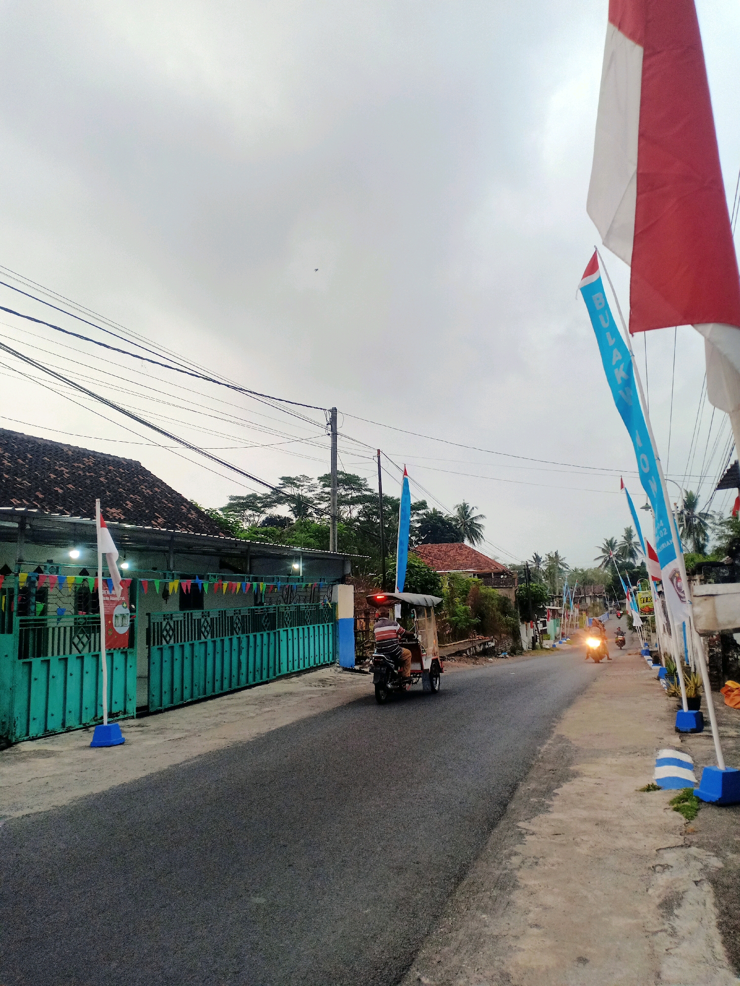 The image depicts a street scene in Pasirian, Indonesia, likely on an overcast day. In the foreground, a paved road runs from left to right, bordered by a sidewalk and vegetation. The left side of the road features a turquoise metal fence in front of a building with a tiled roof and decorative flags. Several power lines are visible overhead.
To the right, the sidewalk is lined with red and white Indonesian flags and blue flags with white lettering, mounted on white poles with blue bases. A few motorcycles are seen on the road, one with a rider in a striped shirt is moving away from the camera. Another motorcycle is approaching with its headlights on. In the distance, more buildings and trees are visible, suggesting a semi-urban or rural setting. The sky is predominantly grey, indicating cloudy weather.