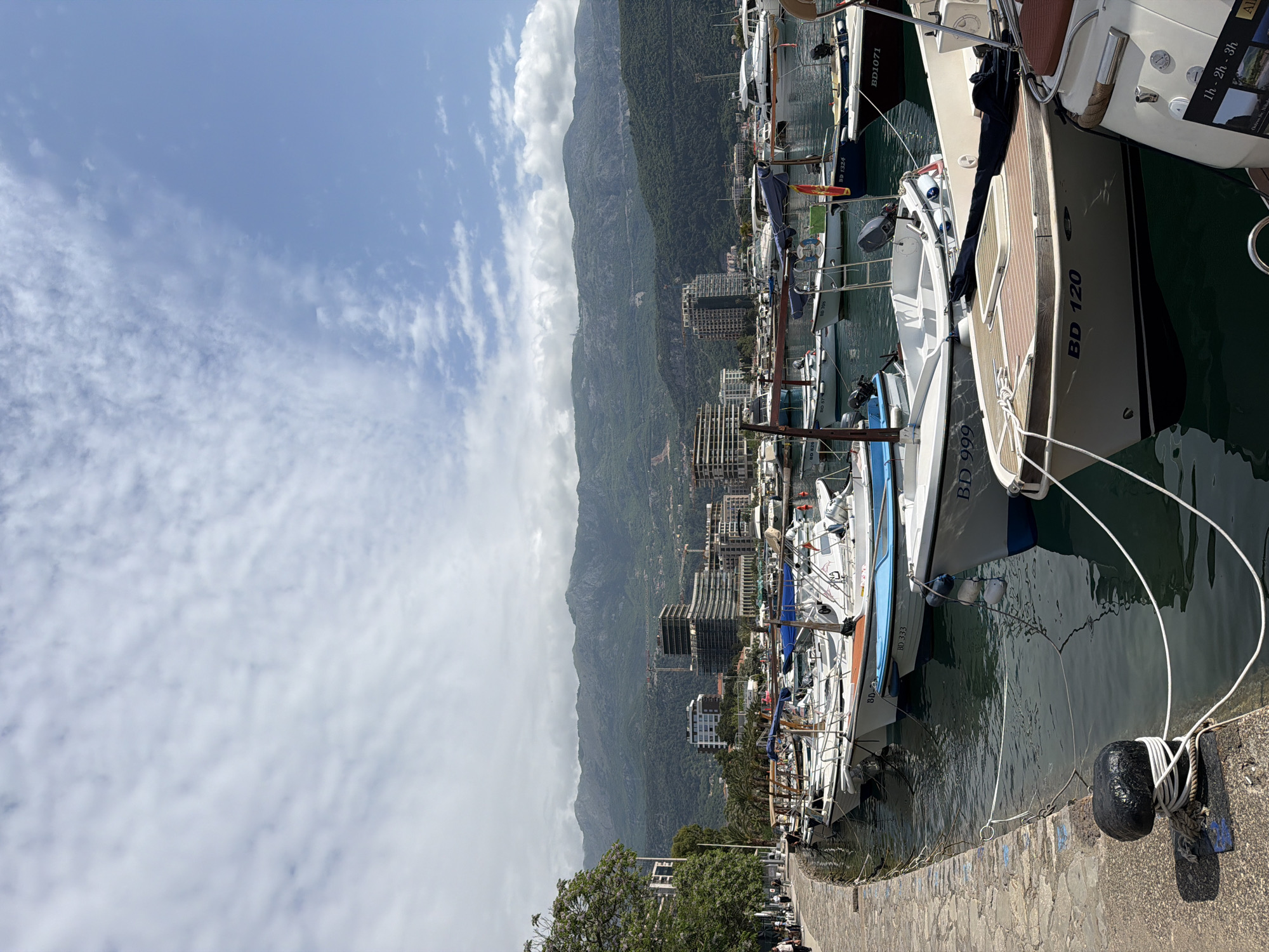 The image depicts a marina scene in Budva, Montenegro, featuring a row of docked boats of various sizes and designs. The closest boat bears the inscription "BD 120" with visible moorings and fenders securing it to the concrete pier. Further along the pier are more boats, including one marked "BD 999." The water in the marina appears calm and reflects the boats and surrounding structures.
In the background, a mountain range rises behind the city's buildings, some of which are under construction. The sky above is partly cloudy.