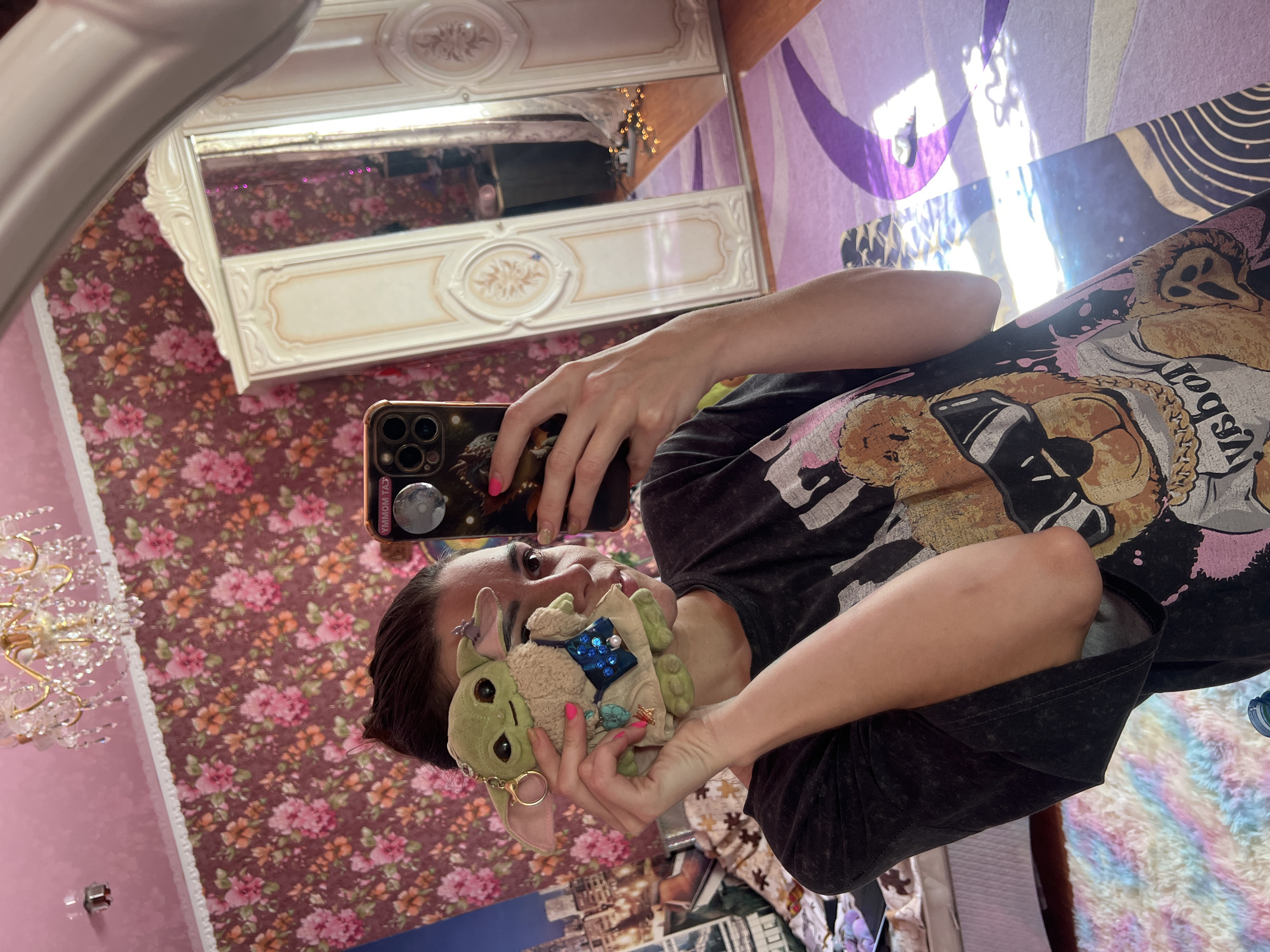 In a bedroom in Krasnoe-na-Volge, Russia, a young woman with dark hair pulled back in a bun takes a mirror selfie. She is holding a Grogu plush toy, a character from Star Wars, in front of her face. The Grogu plush has a tiny blue purse with sequins and a pearl, and a turquoise charm hanging from its neck. The woman is wearing a dark t-shirt with a graphic print of a bear wearing sunglasses.
The room is decorated with floral wallpaper in shades of pink and burgundy. A large white wardrobe with mirrored doors stands to the right of the woman. A chandelier with crystals hangs from the pink ceiling. A purple rug with a swirling pattern covers part of the wooden floor. Sunlight streams in from a window, casting reflections on the rug and wardrobe.