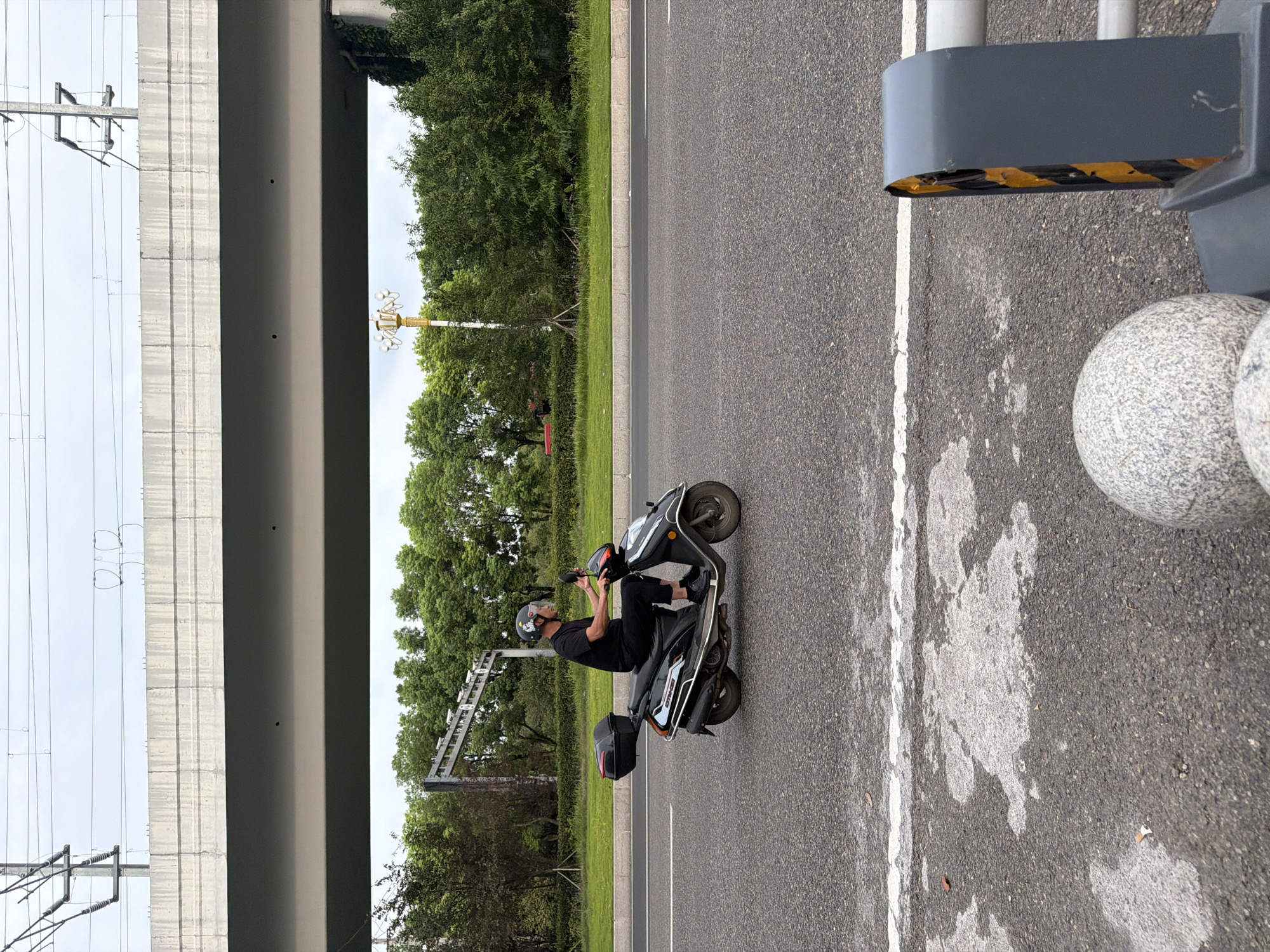 A person wearing a black t-shirt, black pants, black shoes, and a black helmet with a white pattern is riding a black scooter on a paved road. The scooter has a black storage box on the back and a red taillight. The person is looking forward and appears to be in motion. The road is gray and has white lane markings. In the background, there are green trees, a concrete overpass, and utility poles with wires. The sky is overcast. The image was taken in Jiangjin, China.