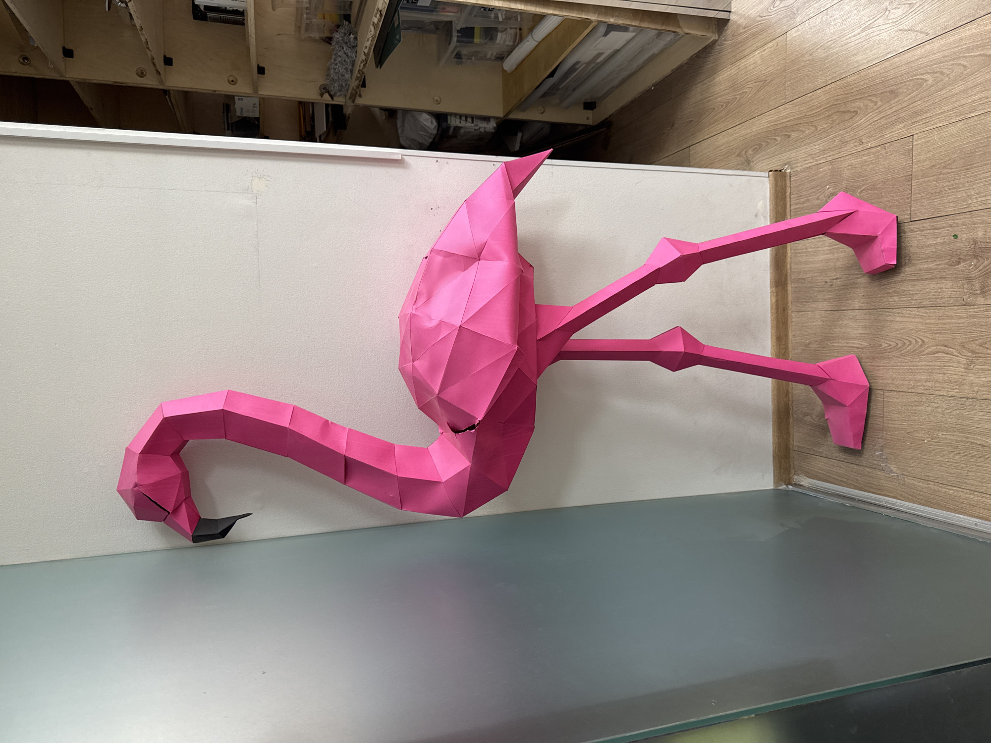A pink paper flamingo with a black beak is affixed to a wall and a glass-topped surface. The wall is light-colored. The surface appears to be a table or countertop. A portion of wooden flooring or paneling is visible on the right side. Overhead, there is a view of an underside, perhaps of a desk or shelf, revealing various electronic components and other items. The location is unknown and cannot be determined from this image. The city of Moscow, Russia, is not visible in the image.