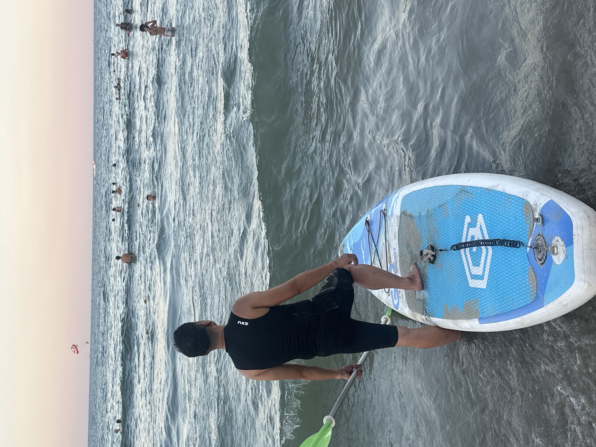 The image shows a man in a black sleeveless shirt and black shorts on a paddleboard. He is holding a paddle with a green blade, and his left foot is on the paddleboard. The paddleboard is blue and white with a textured grip. The man is in shallow water, and waves are visible. In the background, there are several other people in the water, some swimming and some on what appear to be smaller surfboards. Further in the distance, there's a person parasailing. The sky is a soft pink and orange, suggesting it is either sunrise or sunset. The water is calm with some ripples. The scene appears to be a beach in Đà Nẵng, Vietnam. The text "2XU" is visible on the man's shirt. The overall mood is peaceful and recreational.