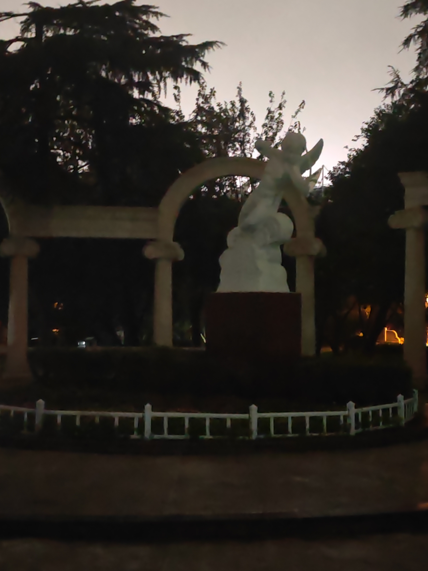 The image captures an outdoor scene at dusk or night, featuring a white statue of a winged figure, possibly an angel, positioned within an arched structure supported by columns. The statue is placed on a dark pedestal. A white, low-lying fence encircles a grassy area in front of the statue. In the background, trees with dark foliage are visible against a light sky. Some artificial lights can be seen illuminating the background, suggesting an urban or park setting. The overall impression is one of a dimly lit, serene monument in a public space. The location is identified as Xuanwu District, China.