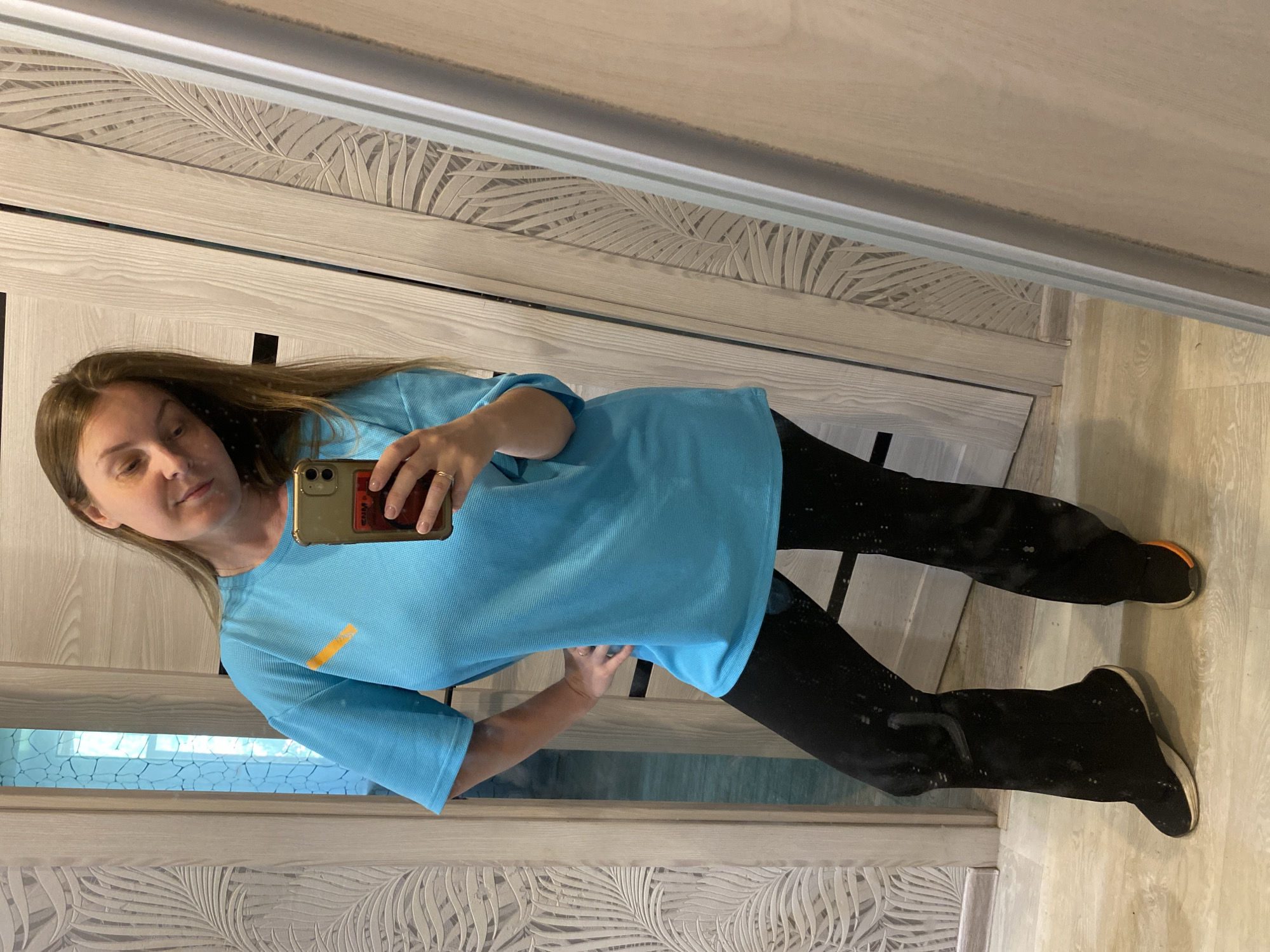 A woman stands indoors, presumably in a room or apartment in Noviy, Russia, based on potential regional indicators. She is taking a selfie in front of a large mirror. She is wearing a light blue t-shirt and black leggings. She is holding a smartphone in her left hand, the screen and camera facing her. Her hair is brown and straight. The mirror reflects a wall with a leaf pattern. The flooring appears to be light wood laminate. The room has wood paneled doors or walls. She is wearing black shoes. There is a thin orange stripe on her shirt.
