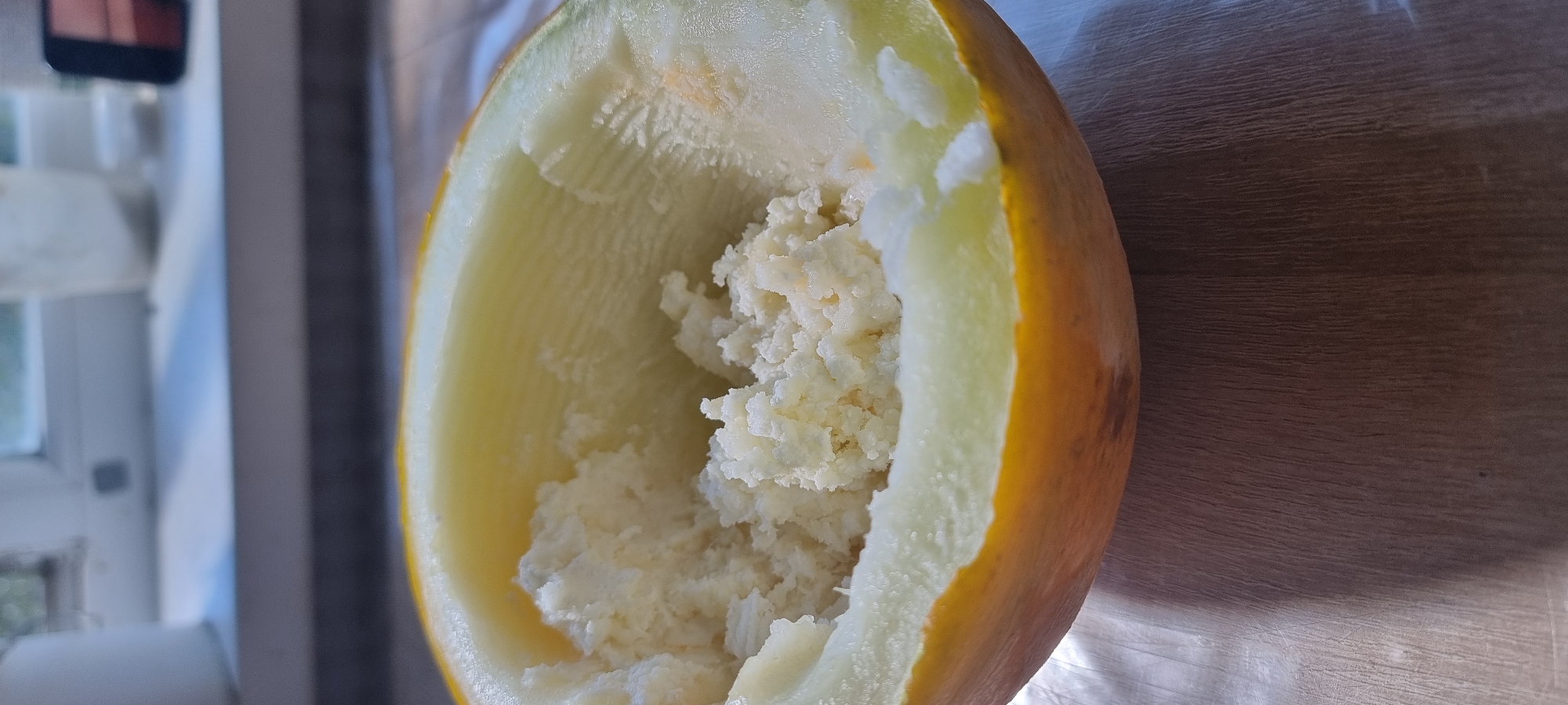 A half-eaten melon, with its flesh scraped out, sits on a wooden surface. The melon is yellow on the outside and pale yellow inside. The background shows a window with sunlight streaming through, illuminating a windowsill with some objects on it, including what appears to be a mobile phone and some containers. The scene is likely indoors, possibly in a residential setting in Aktobe, Kazakhstan, given the context. There are no people or animals visible in the image.