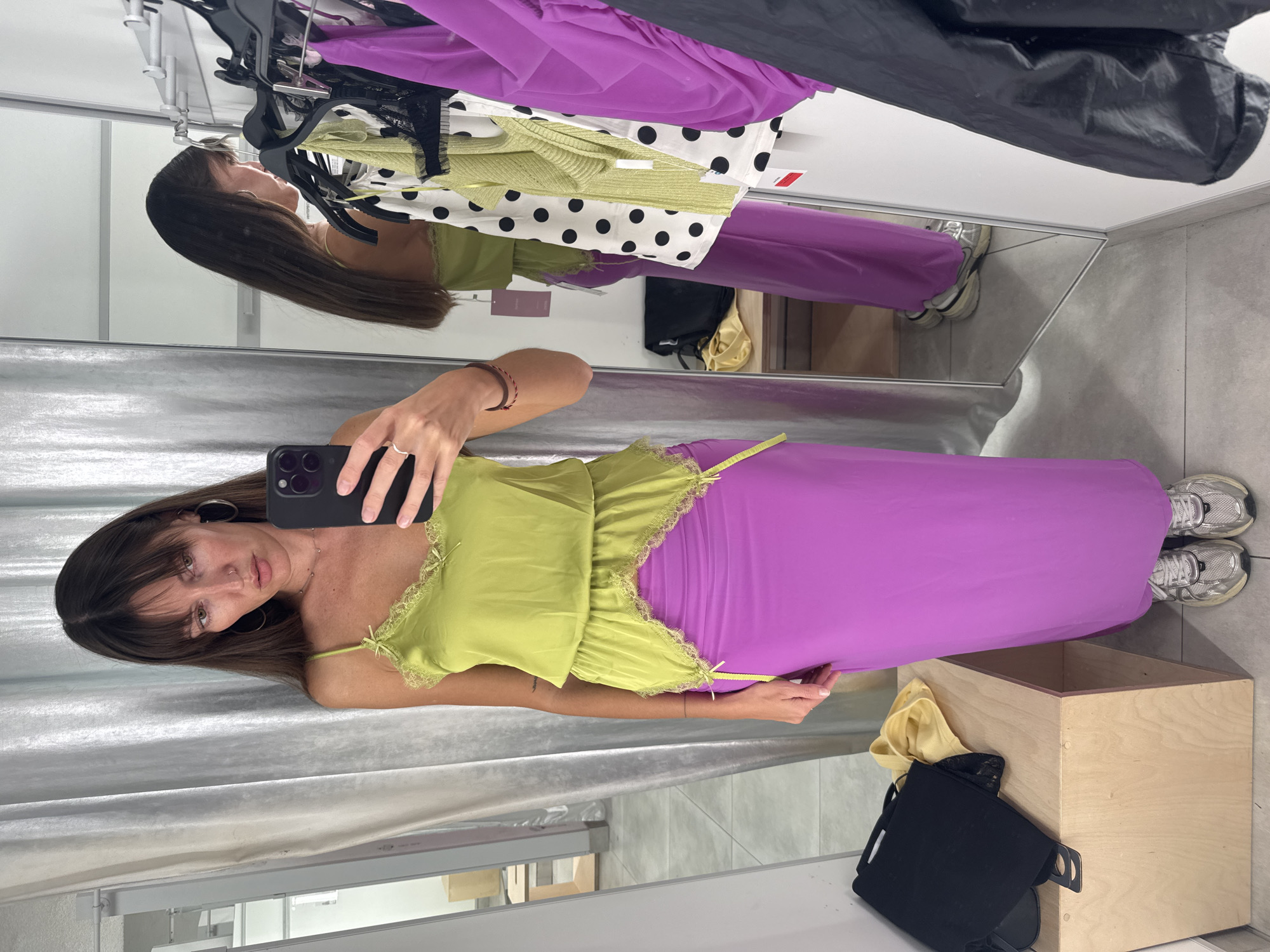 A woman is standing in a fitting room, taking a selfie in a mirror. She is wearing a bright green camisole with lace trim and a long, flowing purple skirt. She has on silver sneakers. In the background, there are clothes on racks, a black bag, and a wooden bench. The fitting room appears to be in a clothing store. The image is taken from a slightly low angle.