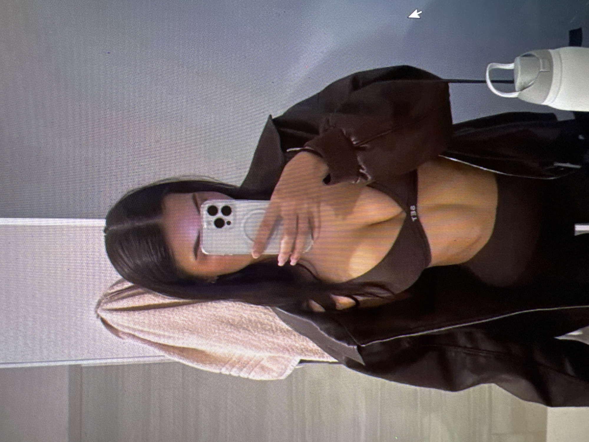 The image shows a woman taking a mirror selfie in what appears to be a bathroom. She is wearing a dark brown bra and a matching set of athletic wear, partially covered by a dark brown jacket. She is holding a smartphone in her right hand, with her face obscured by the phone. A light-colored towel is hanging on a rack to her left, and a white water bottle is visible to her right. The background consists of tiled walls. There is no visible indication of the city of Castelo Branco, Portugal.
