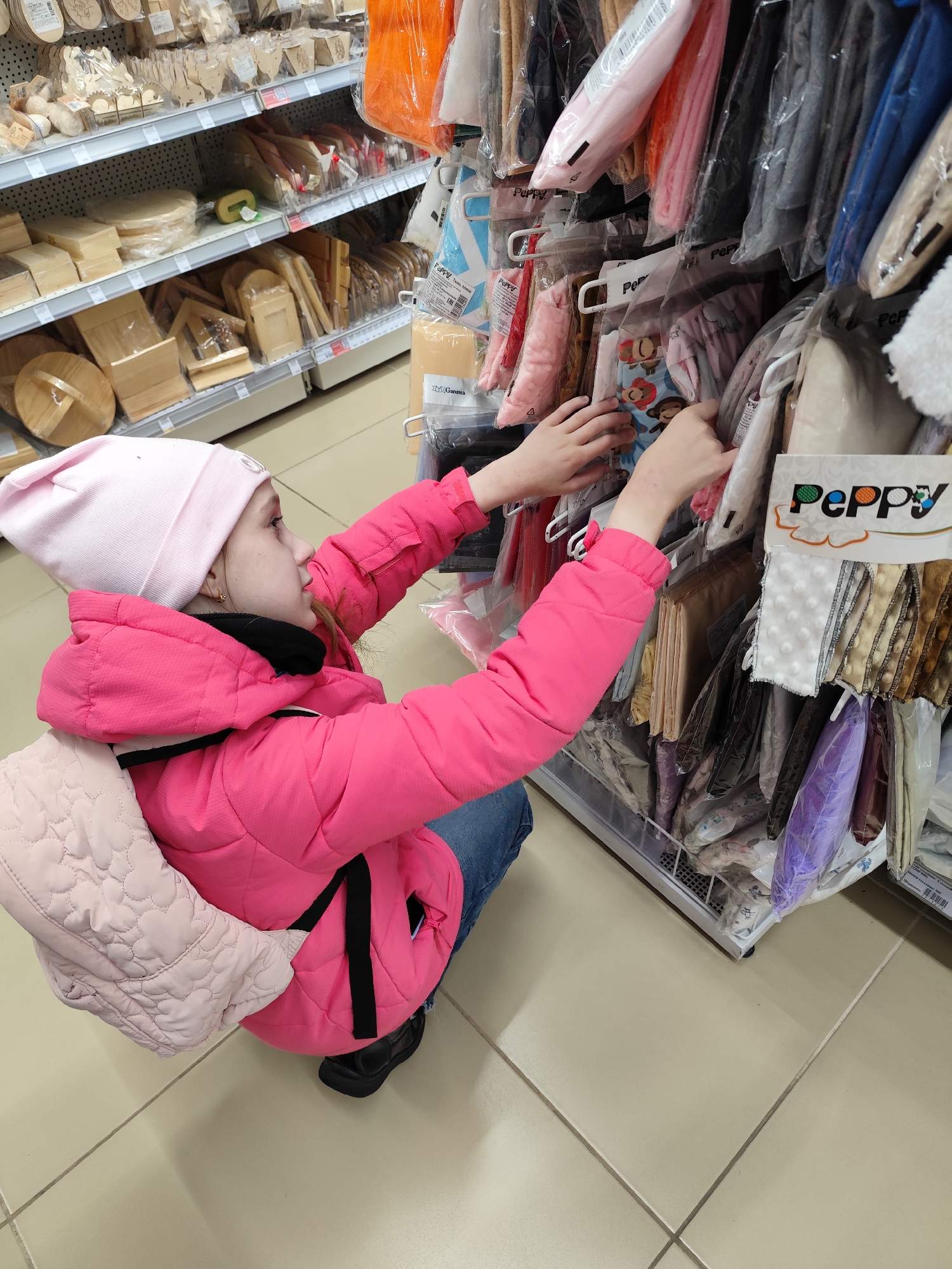 A young girl, viewed from an elevated perspective, crouches down in what appears to be a craft or hobby store, actively examining items on a retail display. She is dressed in a light pink knitted hat, a bright pink puffer jacket with black details, blue jeans, and black shoes. A light-colored, possibly quilted, backpack is on her back.
Her hands are outstretched, sifting through small fabric pieces or craft items, each packaged in clear plastic. These items are hanging from hooks on a metal display rack, with some also stored in a wire basket at the base of the unit. The visible items showcase a variety of colors, including pink, grey, blue, beige, and purple, and some feature patterns, such as one with monkeys. A prominent "Peppy" brand logo is visible on multiple packages and on a display card that also features samples of different fabric textures, including a white fabric with raised dots. The brand name "Gamma" is also faintly visible on another package.
To the left of the girl, in the background, multiple shelves are stocked with various wooden craft supplies, such as wooden cutouts, small boxes, frames, and other unfinished wooden objects. The store floor is covered with light-colored tiles. The indoor lighting does not indicate a specific time of day. The girl appears focused and engaged in the process of selecting an item.