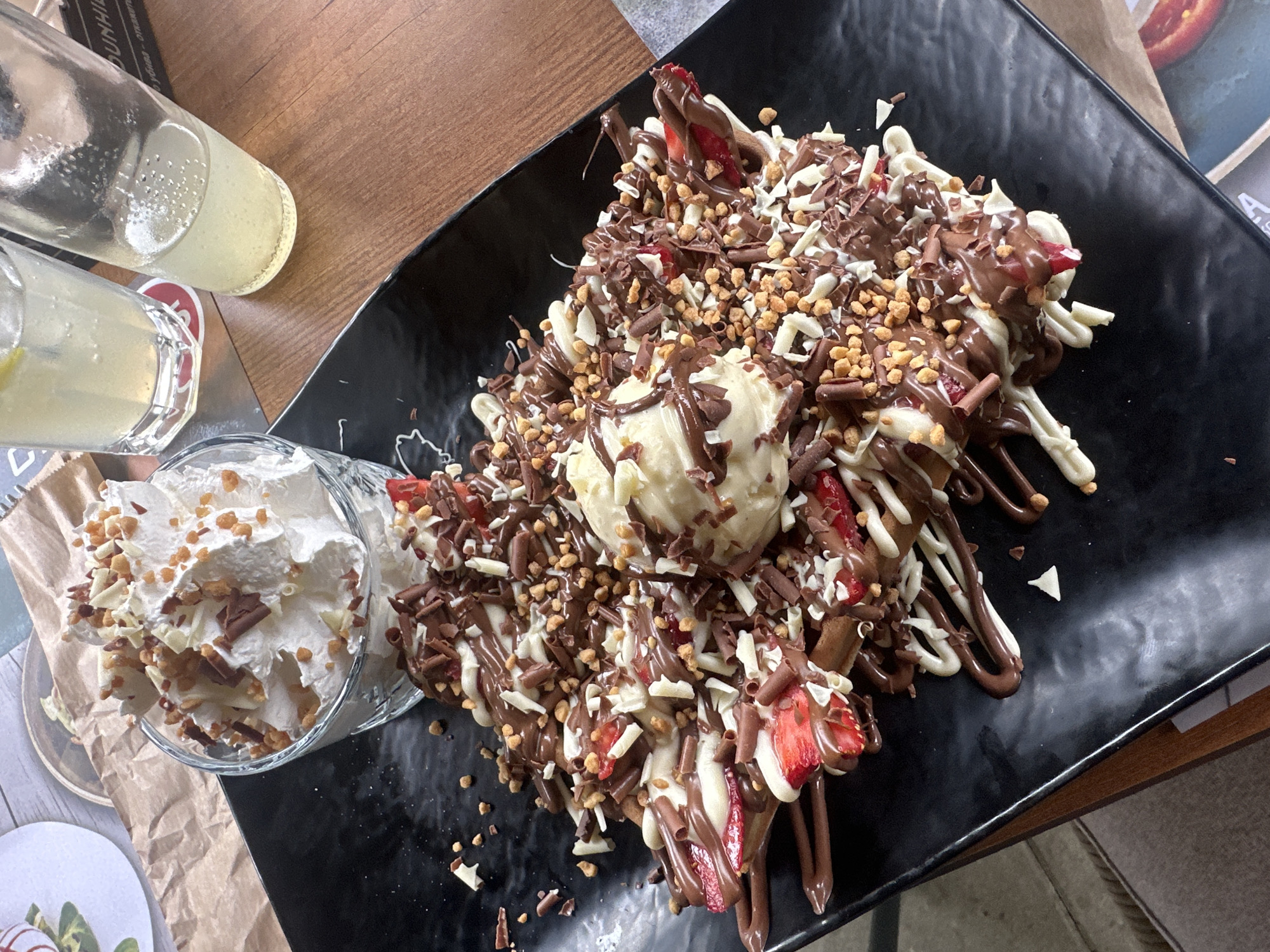 This image captures an overhead view of a large, elaborate dessert and accompanying drinks served on a wooden table within a cafe or dessert parlor in Sofia, Bulgaria.
The primary subject is a substantial dessert presented on a black, rectangular, textured plate. It appears to be a waffle or pancake base, covered with multiple slices of fresh red strawberries. The entire surface is heavily drizzled with both milk chocolate sauce and white chocolate sauce in an alternating pattern. Centrally placed on the dessert is a scoop of light-colored ice cream, likely vanilla. The dessert is generously garnished with an abundance of dark chocolate shavings, white chocolate shavings, and small, light brown granular crumbles, possibly crushed nuts or biscuit pieces.
To the upper left of the main dessert, a small, clear glass bowl holds a large portion of whipped cream. This whipped cream is also topped with the same dark and white chocolate shavings and light brown granular crumbles.
In the upper right portion of the image, two clear glasses or bottles are visible, containing a light yellow, carbonated beverage, presumably lemonade, with bubbles evident. A slice of lemon is visible in the leftmost drink.
The background consists of a medium-brown wooden table with a visible grain. Several paper items are partially visible on the table: a crumpled brown paper bag or liner to the left, and a white paper with partially visible Cyrillic text that includes "бързо - вкусно" (meaning "fast - delicious") under one of the drinks. Beneath the drinks, a black rectangular object, possibly a menu holder or stand, clearly displays the brand name "DUNHILL" in white capital letters, with more partial Cyrillic text underneath it, reading "убива - откето". A dark-colored, possibly upholstered, seating area is faintly visible along the far left edge of the frame. No individuals are visible in the scene. The lighting is bright and even, indicating an indoor setting.