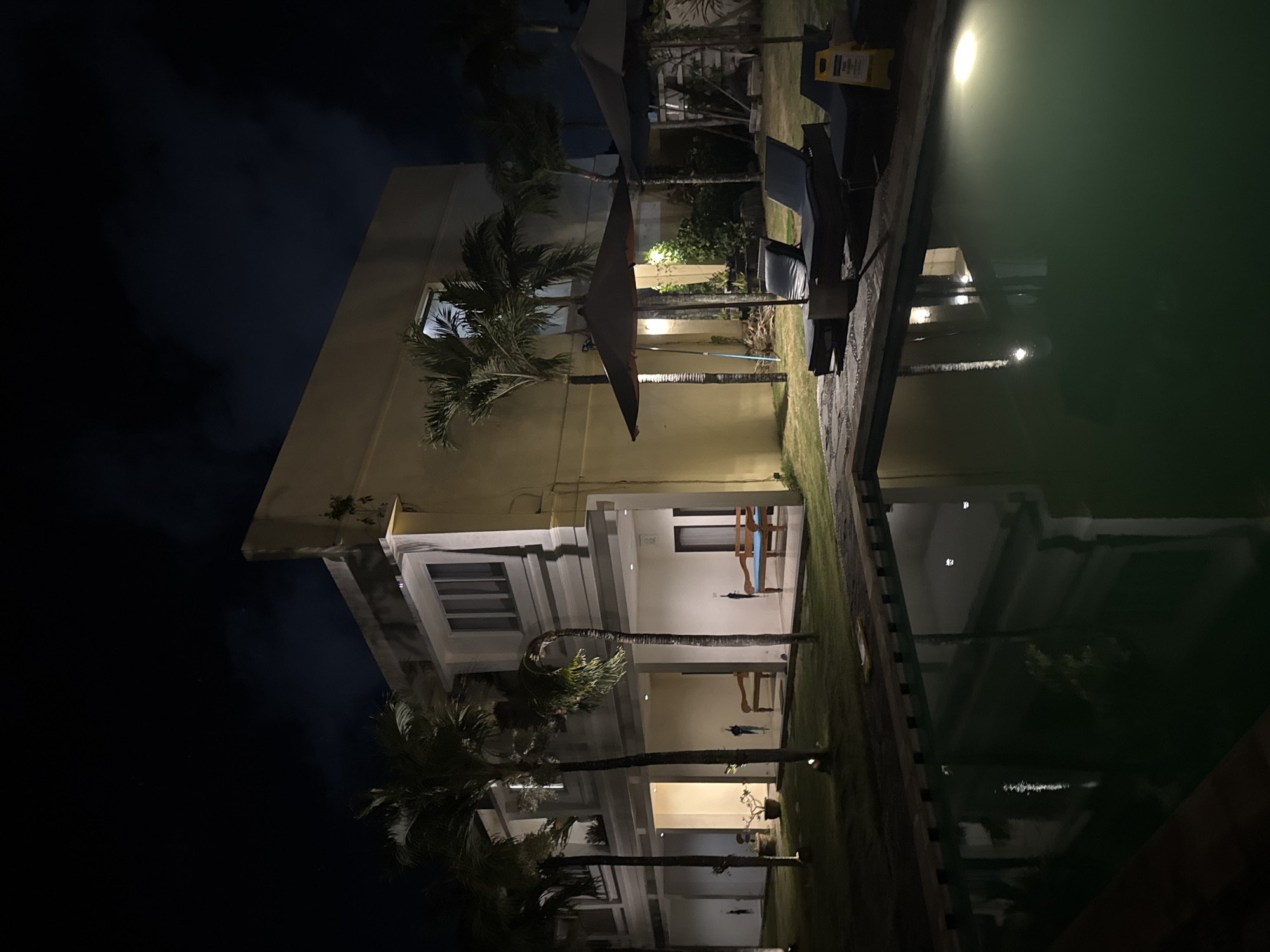 This media file depicts an outdoor scene at night, likely in a tropical location given the presence of palm trees. The setting appears to be a resort or hotel, with a building characterized by light-colored walls and multiple stories. Windows are visible on the building, some illuminated from within, suggesting occupancy.
In the foreground, a dark body of water, likely a swimming pool, dominates the right side of the frame. The water reflects the lights from the building and the surrounding environment, creating a shimmering effect. Along the edge of the water, there are lounge chairs, some of which are empty, hinting at a relaxed atmosphere. Umbrellas are also present, some closed and some open, providing shade during the day and perhaps protection from light rain.
The ground is covered with green grass and paved pathways. The overall lighting is dim, characteristic of nighttime, with artificial lights illuminating specific areas of the building and the pool. The sky is dark, with no visible stars or moon, suggesting it might be overcast or very early/late in the night.
There is no visible text in the media file. The scene evokes a sense of tranquility and relaxation, typical of a holiday destination at night. The orientation of the image is rotated, with the pool appearing to be at the top and the building at the bottom.
