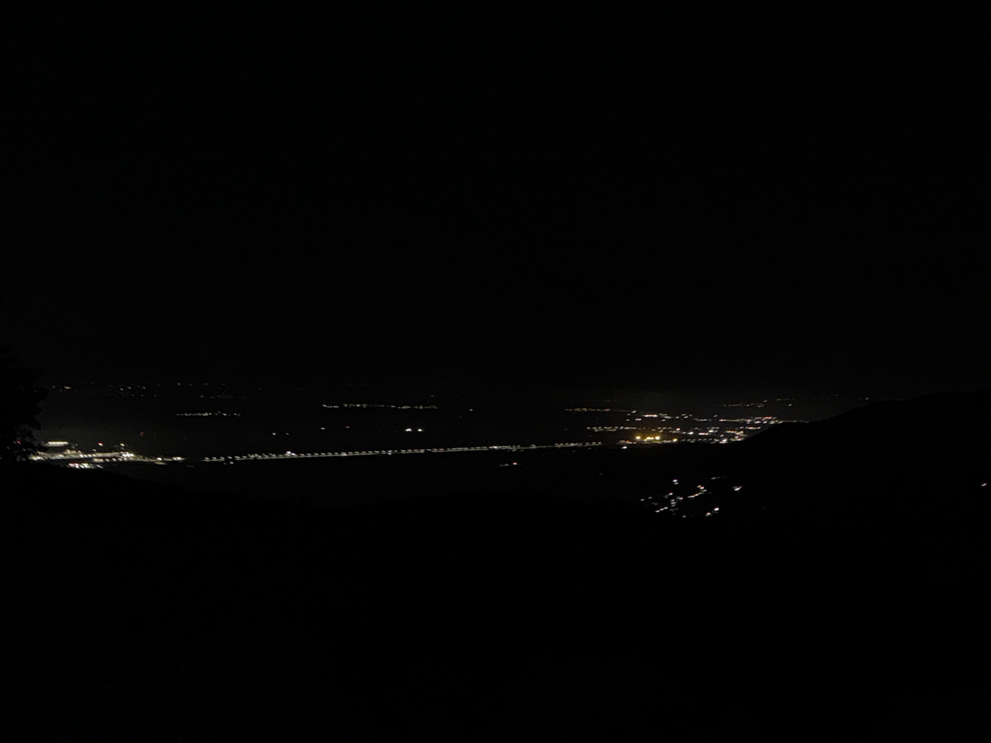 The image presents a broad, dark nighttime landscape dominated by the distant lights of a town or city, viewed from a high vantage point, likely a hill or mountain. The upper half of the frame is almost entirely black, signifying the night sky, with no stars or moon visible.
The main subjects are the numerous artificial lights scattered across the lower half of the image. These lights appear as small, luminous specks, varying in brightness and color, predominantly yellow and white. They form distinct patterns, outlining the extent of human settlement. A prominent linear cluster of lights stretches horizontally across the mid-ground, possibly indicating a major road, railway, or an illuminated feature like a riverbank. To the left, a more concentrated and brighter area of lights suggests a bustling part of the town or a specific facility. On the right, groups of lights ascend gently, hinting at development on sloping terrain.
The foreground is cloaked in deep shadow, with only faint, indistinct dark shapes suggesting the presence of foliage or uneven terrain, from which the photograph was taken. Similarly, the areas beyond the illuminated settlement are submerged in darkness, with subtle dark outlines in the far distance indicating further hills or mountains.
The time of day is clearly night. The air appears mostly clear, allowing the distant lights to be seen distinctly, though there is a slight atmospheric haze. No specific activity or event is discernible beyond the static display of urban illumination. No people or identifiable objects other than the lights and the dark landscape are visible. There is no visible text. The scene conveys a sense of peaceful vastness and the quiet glow of civilization against the natural darkness. Given the context, this is a night view of Galabovo, Bulgaria, or its immediate surroundings, from an elevated perspective.