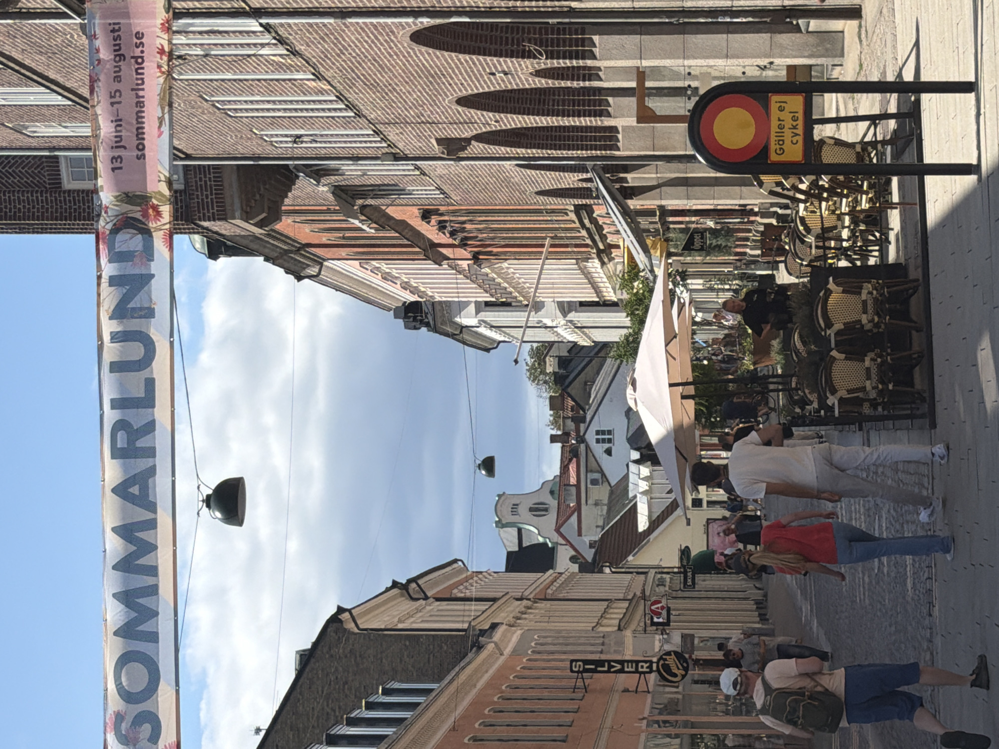 The image depicts a street scene in Lund, Sweden, likely during the summer festival "Sommarlund," as indicated by a banner across the street that reads "SOMMARLUND." The banner also includes dates "13 juni - 15 augusti" and a website "sommarlund.se." The street is paved with cobblestones and lined with buildings. On the left, there are buildings with light-colored facades and awnings, and signage for "SILVER" and "Guild." Further down the street, more buildings are visible, including one with a white facade and decorative elements. On the right side of the street, buildings are constructed of brick, featuring arched ground-floor openings. A sidewalk cafe with tables and chairs is set up on the right, covered by a large white umbrella. In the foreground, three people are walking away from the camera. A man with a backpack is on the left, followed by a woman in a red top and blue jeans, and a man in a white t-shirt and light-colored pants. A street sign with a red circle and yellow circle, and text "Gäller ej cykel" (Not applicable to bicycles) is prominently displayed on the right side of the image. The sky is blue with some clouds. Various shops and establishments are indicated by signage along the street.