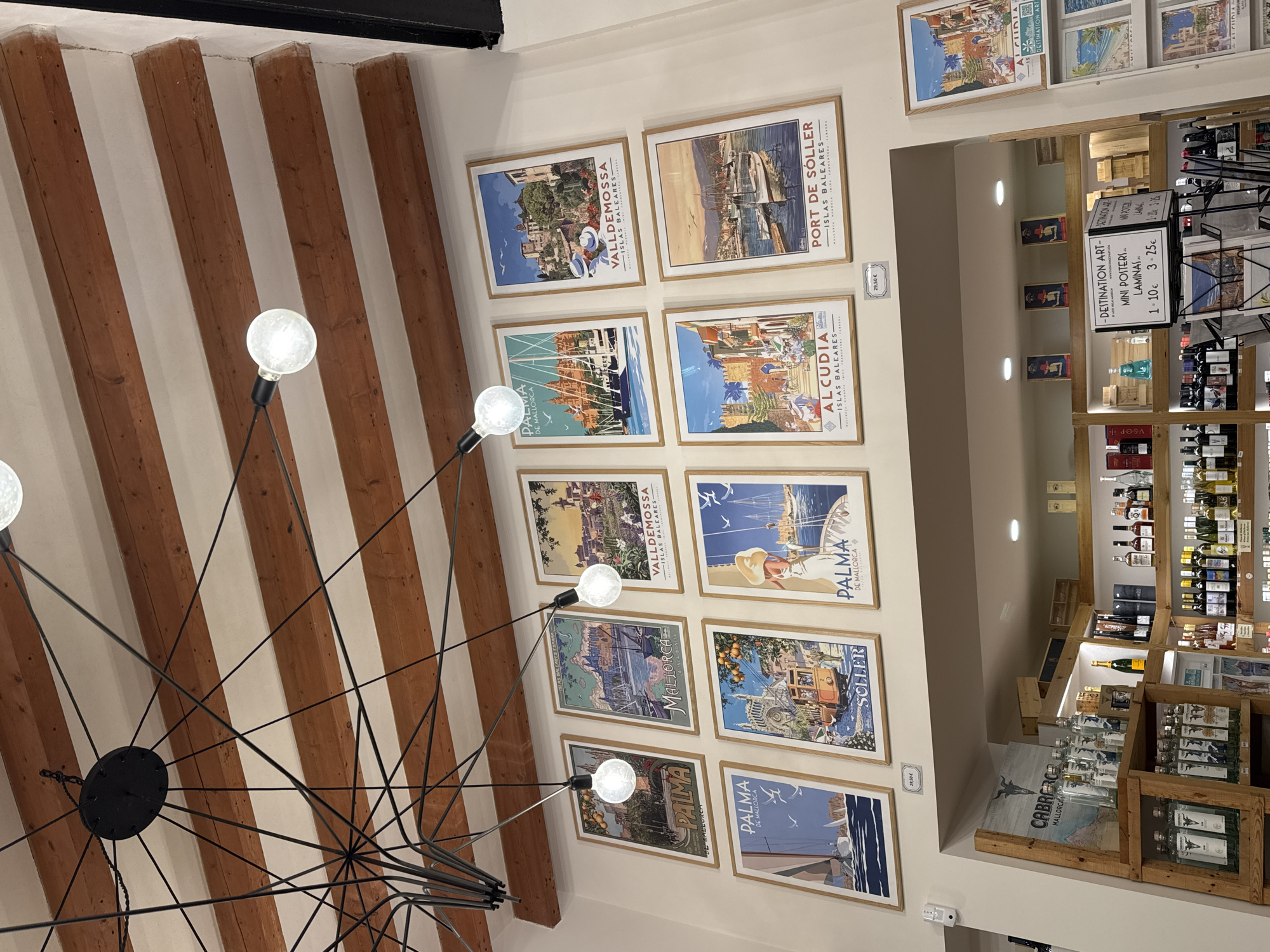 The image captures an interior scene, likely within a retail or commercial space in Alcúdia, Spain. The primary focus is a wall adorned with framed vintage-style travel posters depicting various locations in Mallorca, Spain, including Valldemossa, Port de Sóller, Alcúdia, Palma, and Sóller. The posters are arranged in a grid pattern, suggesting they are for sale.
To the right, a shelving unit holds a variety of products, including what appears to be wine, spirits, and other retail items. A small sign displays pricing for "Destination Art" mini posters and laminas, suggesting the location is a tourist shop.
Overhead, exposed wooden beams support the ceiling, and a modern chandelier with multiple exposed bulbs hangs prominently in the foreground.
The overall impression is that of a tourist-oriented shop selling local art and products related to Mallorca. The scene conveys a sense of place, showcasing the charm and appeal of the island as a travel destination.