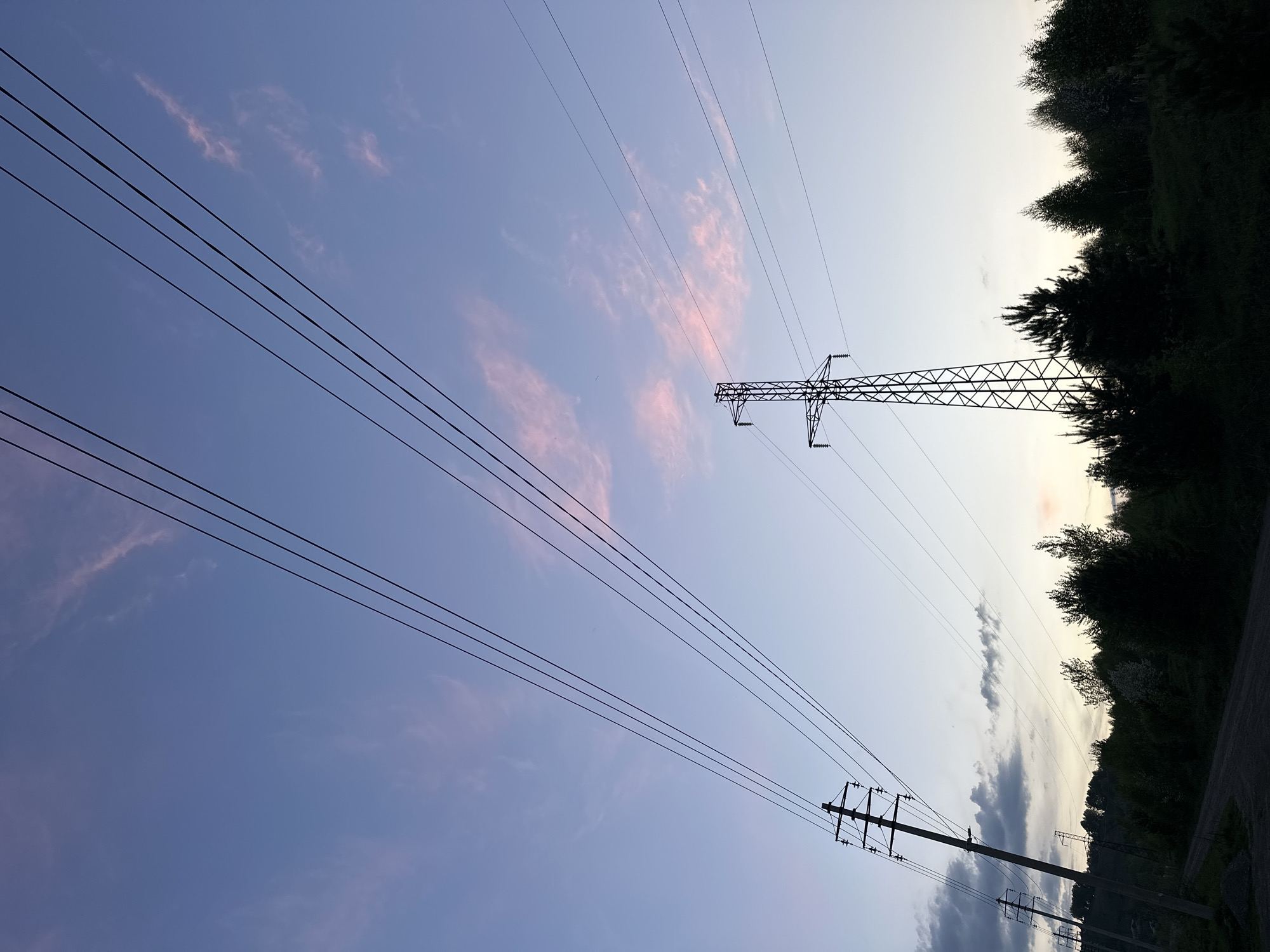 The image shows a low-angle view of the sky with power lines running across it. The sky is light blue with streaks of pink clouds. Two power line towers are visible, one larger and more complex than the other. The edges of a forest are visible in the upper right of the frame, appearing dark against the sky. The lower right of the image reveals a small portion of a road, suggesting the scene is likely from a location within or near Gorno-Altaysk, Russia, potentially on the city outskirts or in a residential area with power lines running through it. There are no people visible in the frame.