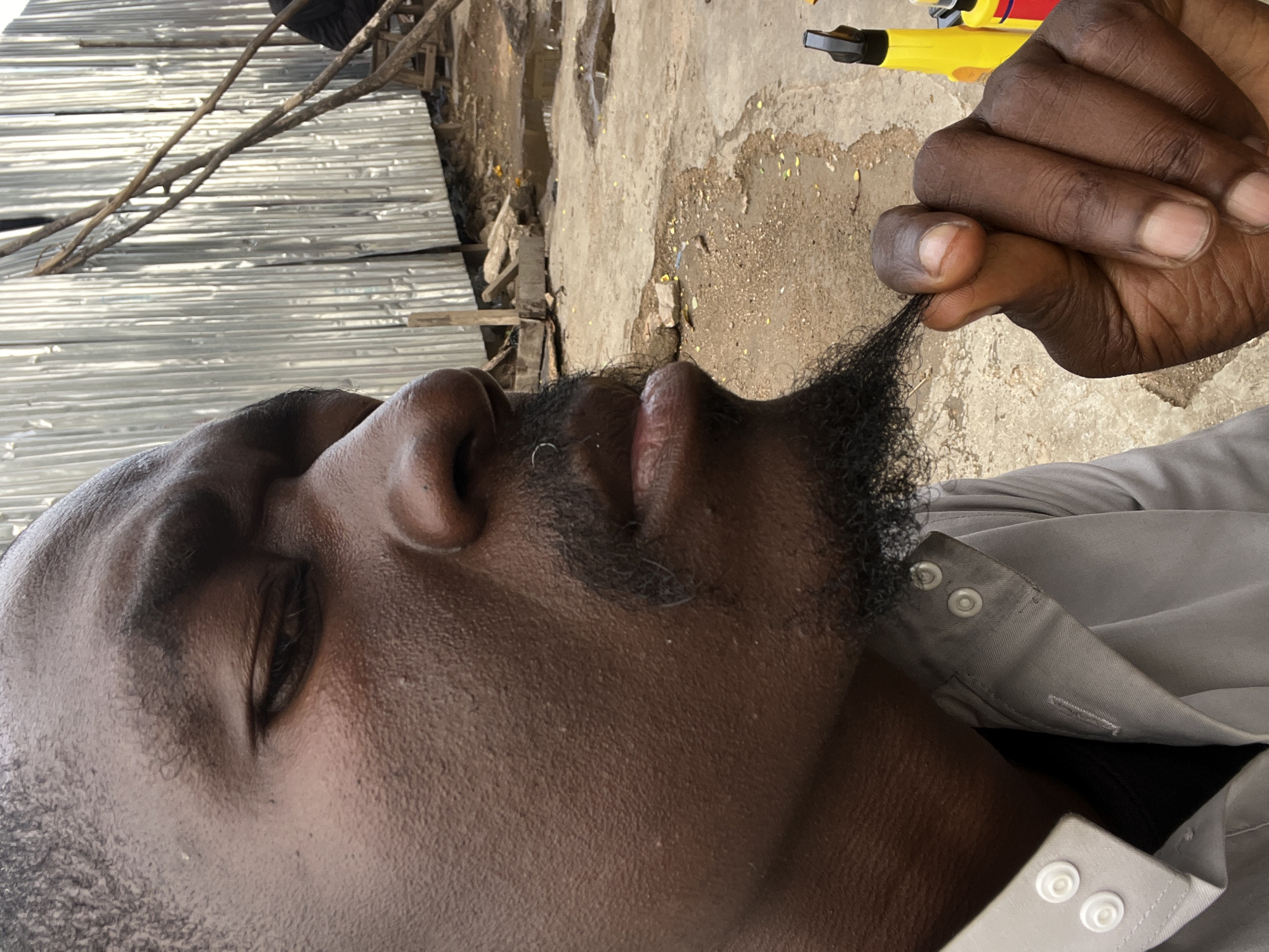 A close-up outdoor shot, rotated ninety degrees clockwise, features a dark-skinned person from the chest up in Jos, Nigeria. The person's eyes are closed, and their right hand is prominently displayed, gently pinching a small section of their dark, short beard near their chin. A small, circular, silver-colored nose ring is visible on their right nostril. They wear a light grey collared shirt with white snap buttons visible on the placket.
The background, viewed as the top of the image in its original orientation, features corrugated metal sheets on the far left and rustic wooden branches extending across the upper left. The ground on the right side of the background is a mix of cracked concrete and dirt, with scattered light debris or leaves. Part of a yellow and black object, possibly a tool, is partially visible in the upper right corner. The clear lighting suggests it is daytime. The combination of corrugated metal, rough ground, and wooden elements indicates an informal, possibly urban or semi-urban setting. No text is visible in the image.