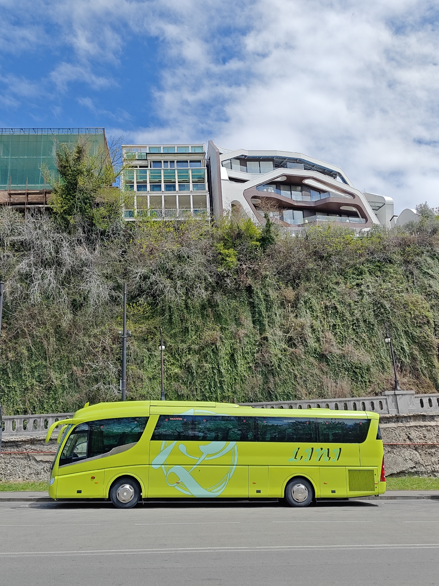 A bright lime green bus with light blue stylized decals is parked on the side of an asphalt road under a clear blue sky with scattered white clouds. The bus features a distinct, aerodynamic design and has "LIJILI" (stylized text) printed in light blue on its rear side, along with swirling patterns further forward.
Behind the bus, a steep hillside rises, densely covered in various shades of green foliage, including ivy and shrubs, with some areas showing bare, interwoven branches. A gray concrete retaining wall with a railing runs along the base of the hill.
On top of the hill, several modern buildings are visible. To the left, a building is partially covered by green scaffolding mesh. In the center, a striking contemporary building stands out with its undulating, organic architectural style, featuring curved brown and white facades that frame large glass windows and balconies. Another modern structure with large windows is visible next to it. The scene is brightly lit, indicating daytime. No individuals are visible. The setting depicts a blend of natural incline with modern urban development.