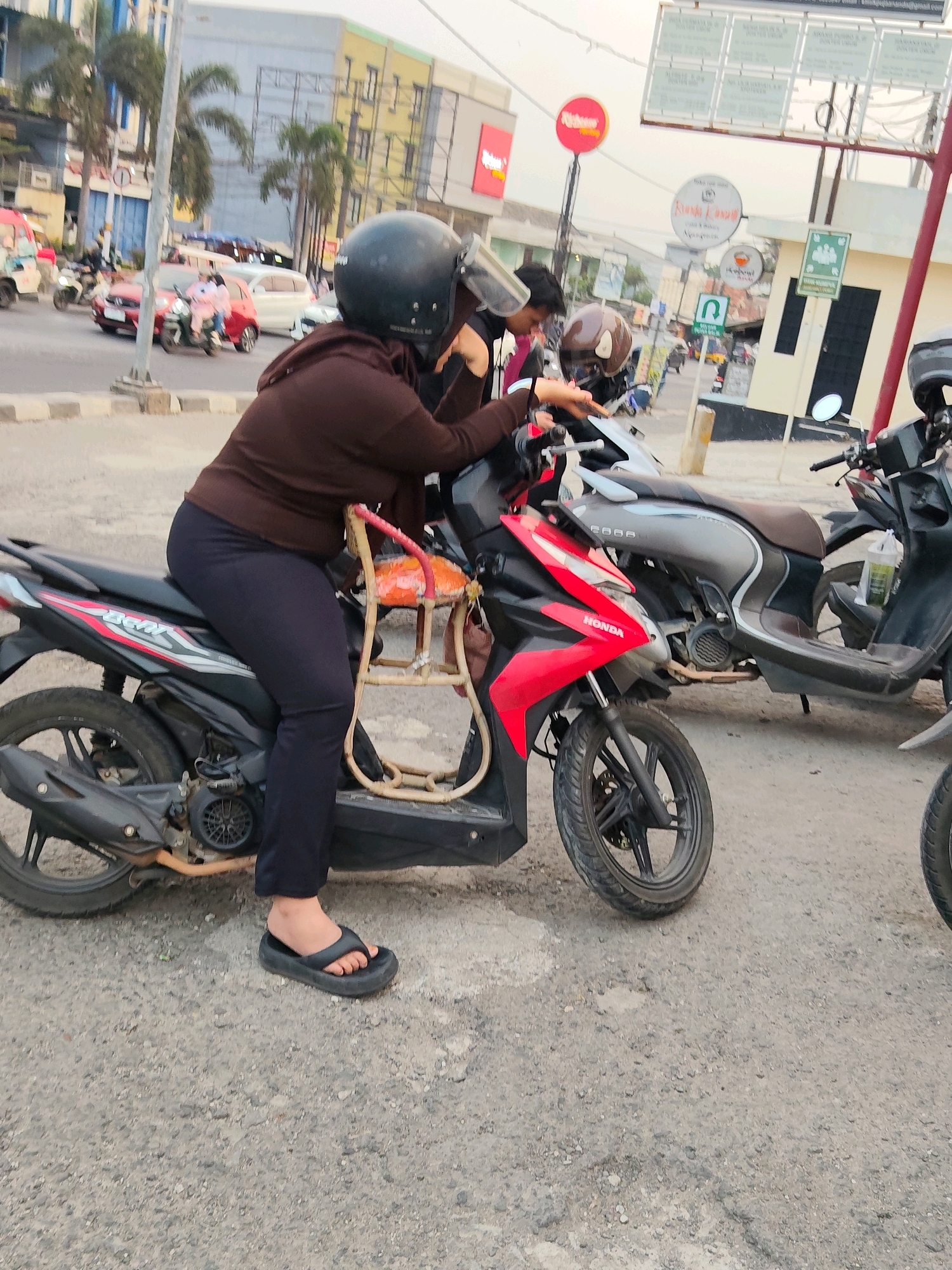 A side view shows a woman wearing a black helmet, a brown hooded sweatshirt, and dark pants, sitting on a red and black Honda motorcycle. She is holding onto the handlebars, and her left foot is on the ground wearing a black flip-flop. A child's seat with an orange cushion is attached to the back of the motorcycle. The motorcycle is parked on a paved street, with other motorcycles and cars visible in the background. In the distance, buildings and street signs are present, suggesting an urban environment in Purwakarta, Indonesia. The overall scene depicts a moment of pause on a street, possibly waiting or preparing to depart.