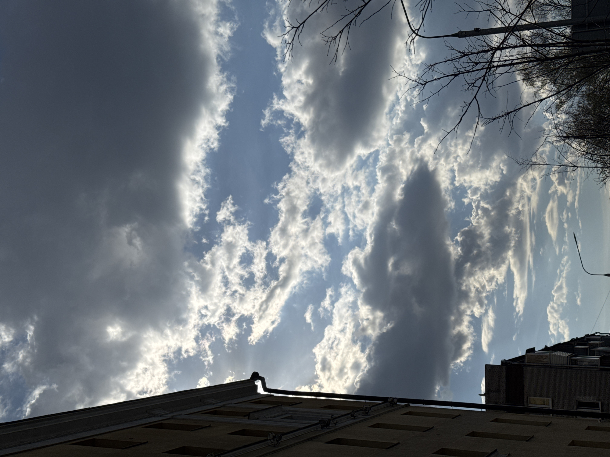 The image shows a view looking upwards at a sky filled with clouds, including dark storm clouds, and an urban environment. The location is Moscow, Russia. The view is from street level, with parts of buildings visible in the foreground. One building has a tan or yellow facade with rectangular features. Bare tree branches and streetlights are visible in the upper right corner of the image.