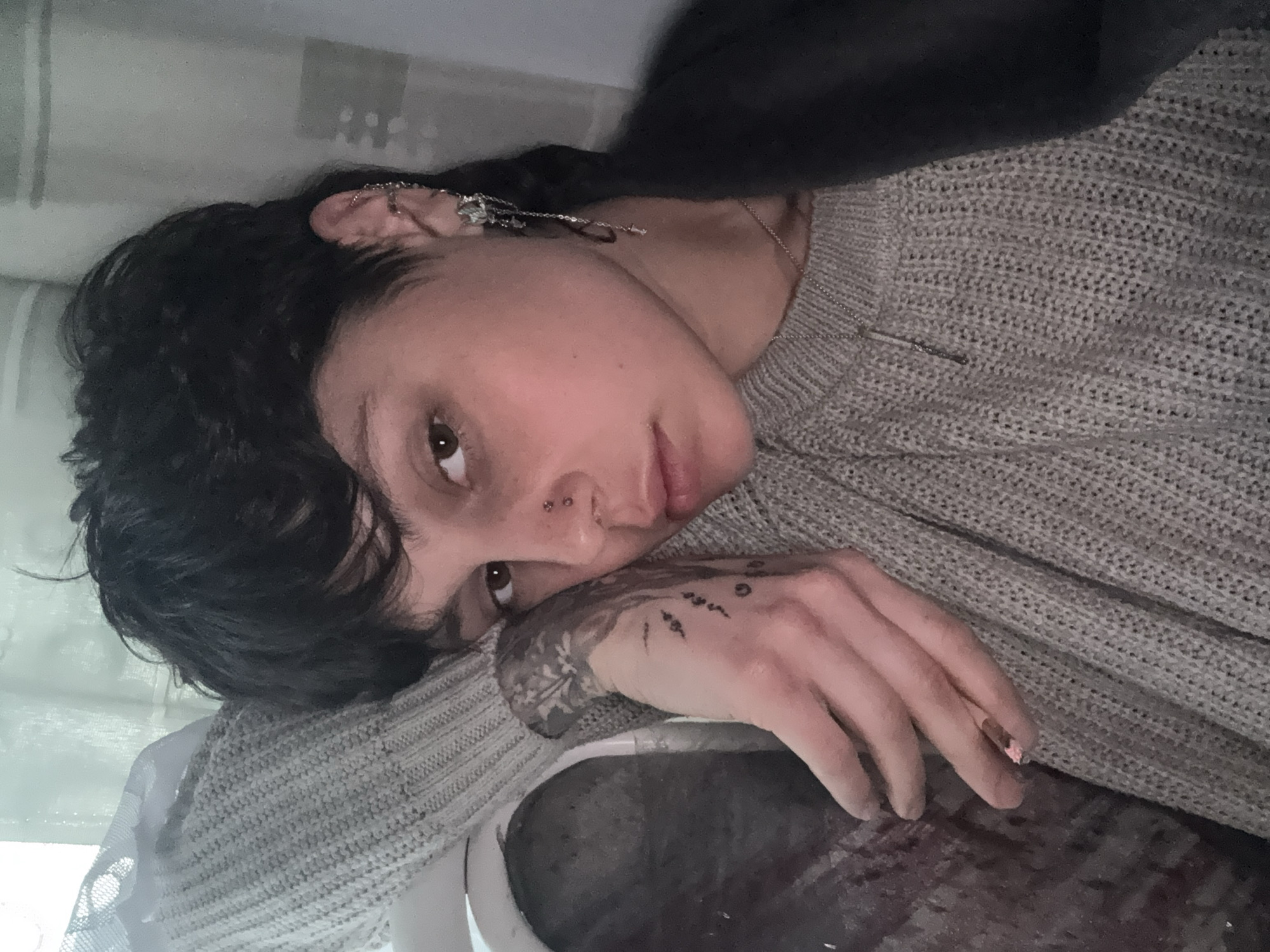 A young person with short, dark, wavy hair and brown eyes is depicted lying down, looking directly at the camera. They have a calm, neutral expression. They are wearing a light grey, knitted sweater with a visible texture.
The person sports multiple ear piercings, including several helix piercings, an industrial bar, and dangling earrings featuring small charms. A double nose piercing with two small studs is visible. A thin, silver-colored necklace with a delicate pendant rests on their collarbone. Intricate tattoos adorn their right forearm, with what appears to be a geometric or abstract design, and smaller symbolic tattoos are visible on their knuckles. A hint of pink, possibly from chipped nail polish, is seen on their right pinky finger.
They are resting their head on their right arm, which is positioned on a dark, textured surface in the foreground, possibly a piece of furniture or a dark blanket. In the background, a soft, light-colored fabric, resembling a curtain or blind, is visible, suggesting an indoor setting with natural light. A dark, blurry shape is present near the top of the frame, possibly part of their clothing or another object. The overall scene suggests a moment of relaxation. No visible text is present in the image. The location context provided is Saint Petersburg, Russia.