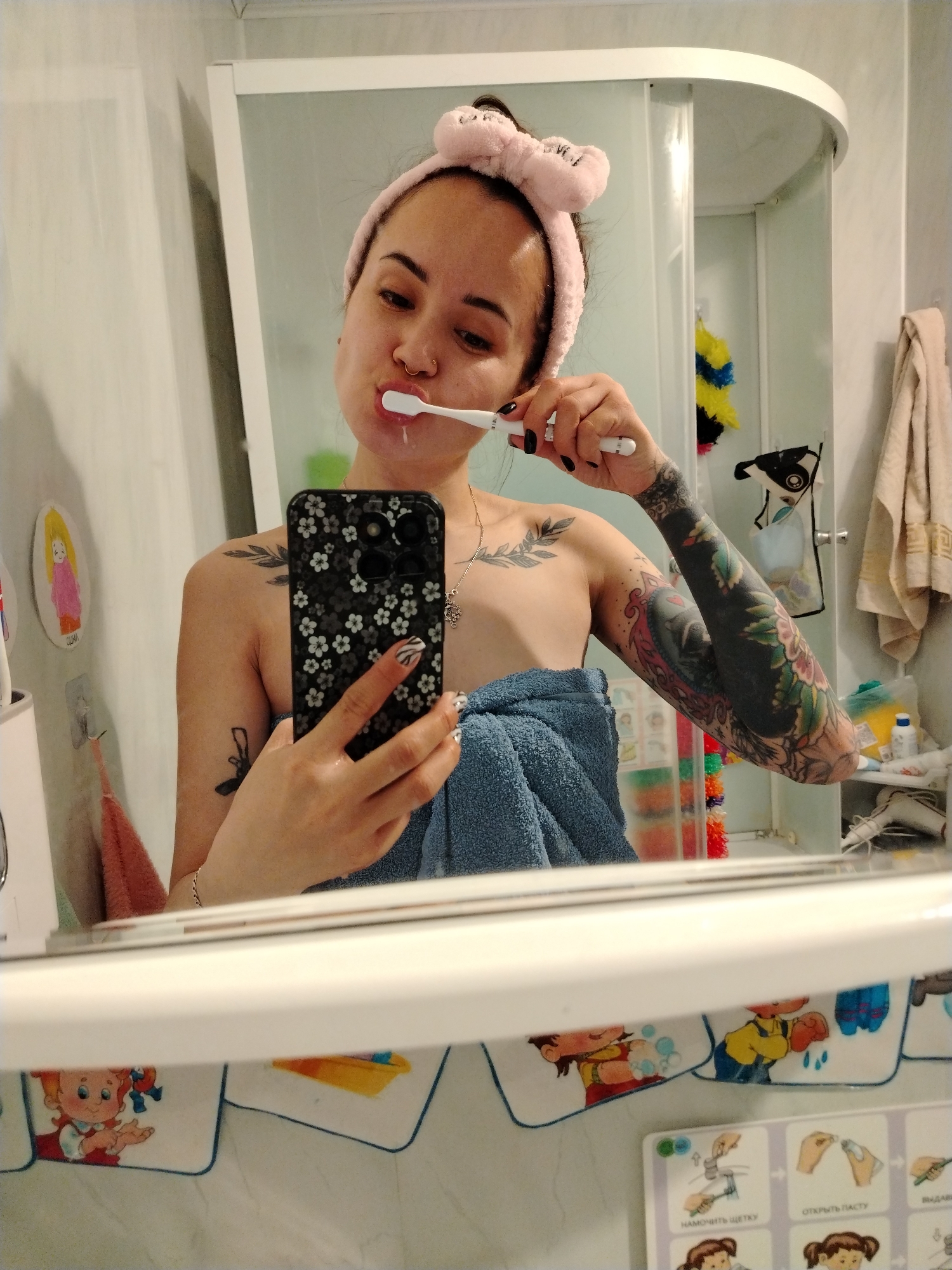 A young woman is captured in a mirror selfie as she brushes her teeth in a bathroom. She is seen from the chest up, with dark hair pulled back and held by a pink, fuzzy headband. A nose ring is visible, and she has several tattoos, including a prominent full sleeve on her left arm, a shoulder tattoo, and another smaller design on her right arm/shoulder area. Her fingernails are painted black, with some featuring a white zebra pattern. She is wrapped in a blue towel.
In her right hand, she holds a white toothbrush, actively brushing, with toothpaste foam visible around her mouth. Her left hand holds a black smartphone with a distinctive white floral pattern case, which she is using to take the photo.
The bathroom setting includes a white-framed shower cabin behind her, containing a yellow and blue bath pouf and some small bottles on a shelf. To the left, a white toothbrush holder and a small cartoon-like magnet (a blonde girl in a yellow oval) are affixed to the wall. A beige towel hangs on a rack to the right. Below the mirror, children's pictorial instructions for hygiene are visible, detailing steps for washing hands and brushing teeth. Visible Russian text on these charts includes "НАМОЧИТЬ ЩЕТКУ" (Wet the brush), "ОТКРЫТЬ ПАСТУ" (Open the paste), and "ВЫДАВИТЬ" (Squeeze out). The scene appears to be illuminated by indoor lighting, suggesting a routine personal moment. This domestic scene is set in Noviy, Russia.