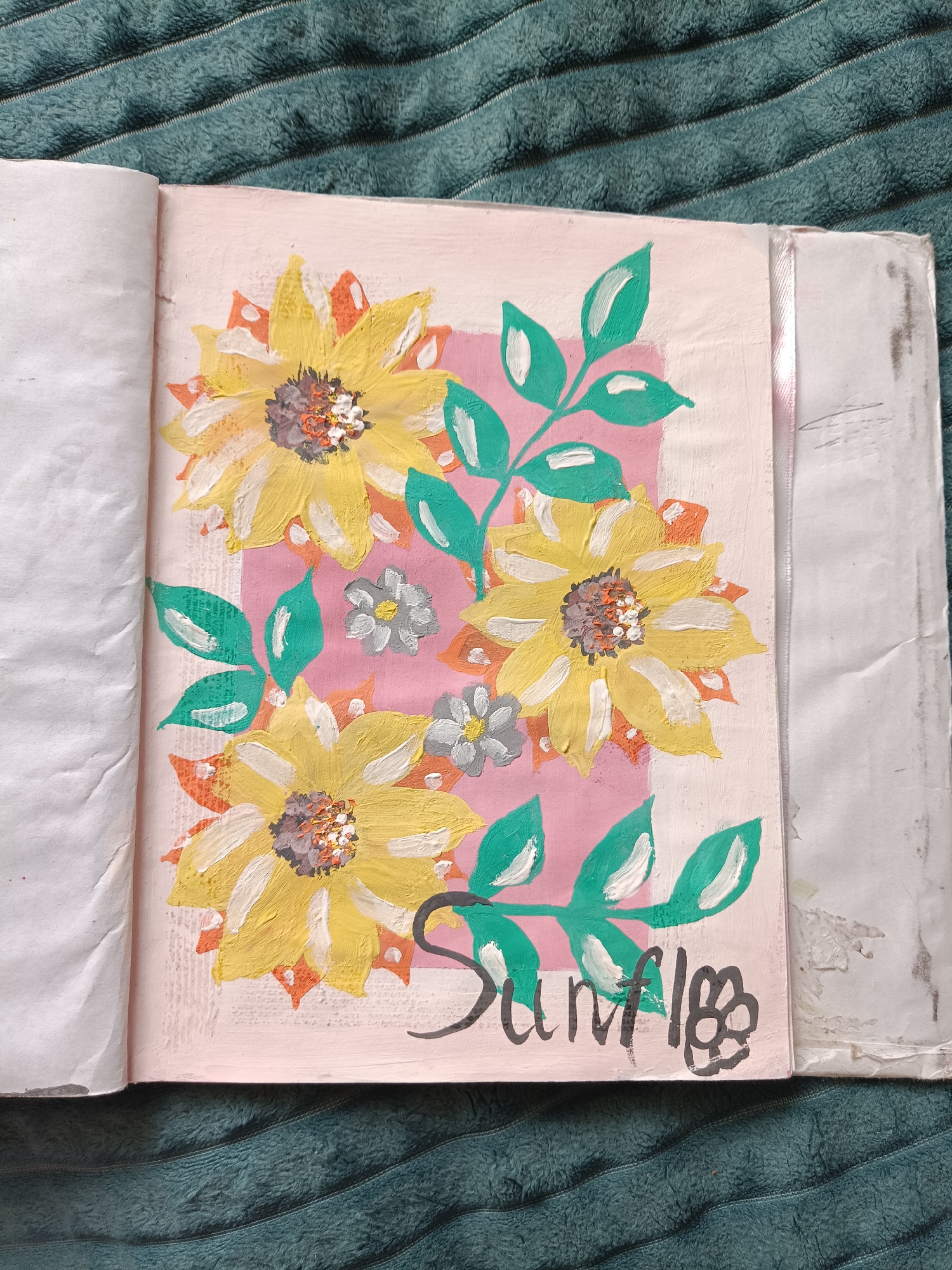 An open scrapbook lies on a textured, teal-colored surface. The right-hand page features a painting of three large sunflowers with green leaves and two smaller, gray flowers. The sunflowers have yellow petals with accents of white and orange, and brown and white centers. The background is a pink rectangle surrounded by a beige-white border. The word "Sunflo" is written in black ink below the flowers, with the letters "wer" being depicted as a stylized flower. The left-hand page is blank and wrinkled. The scrapbook appears worn.
I do not have information about location details related to the city of Посад, Russia.