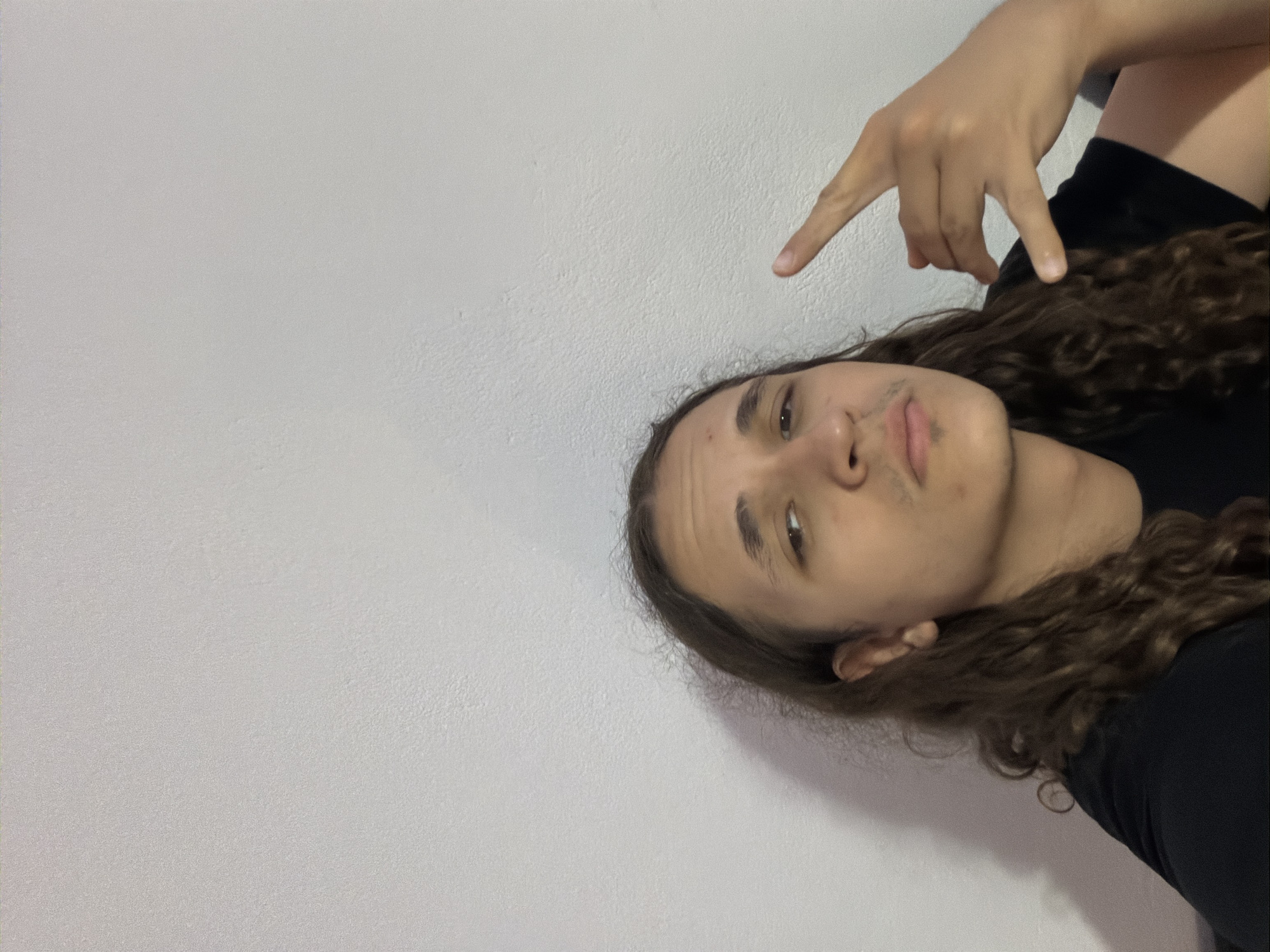 A person with long, curly brown hair is looking at the camera. They are wearing a black shirt. They are pointing upwards with their right hand. The background is a light gray wall. There is no indication of the city of Asenovgrad, Bulgaria.