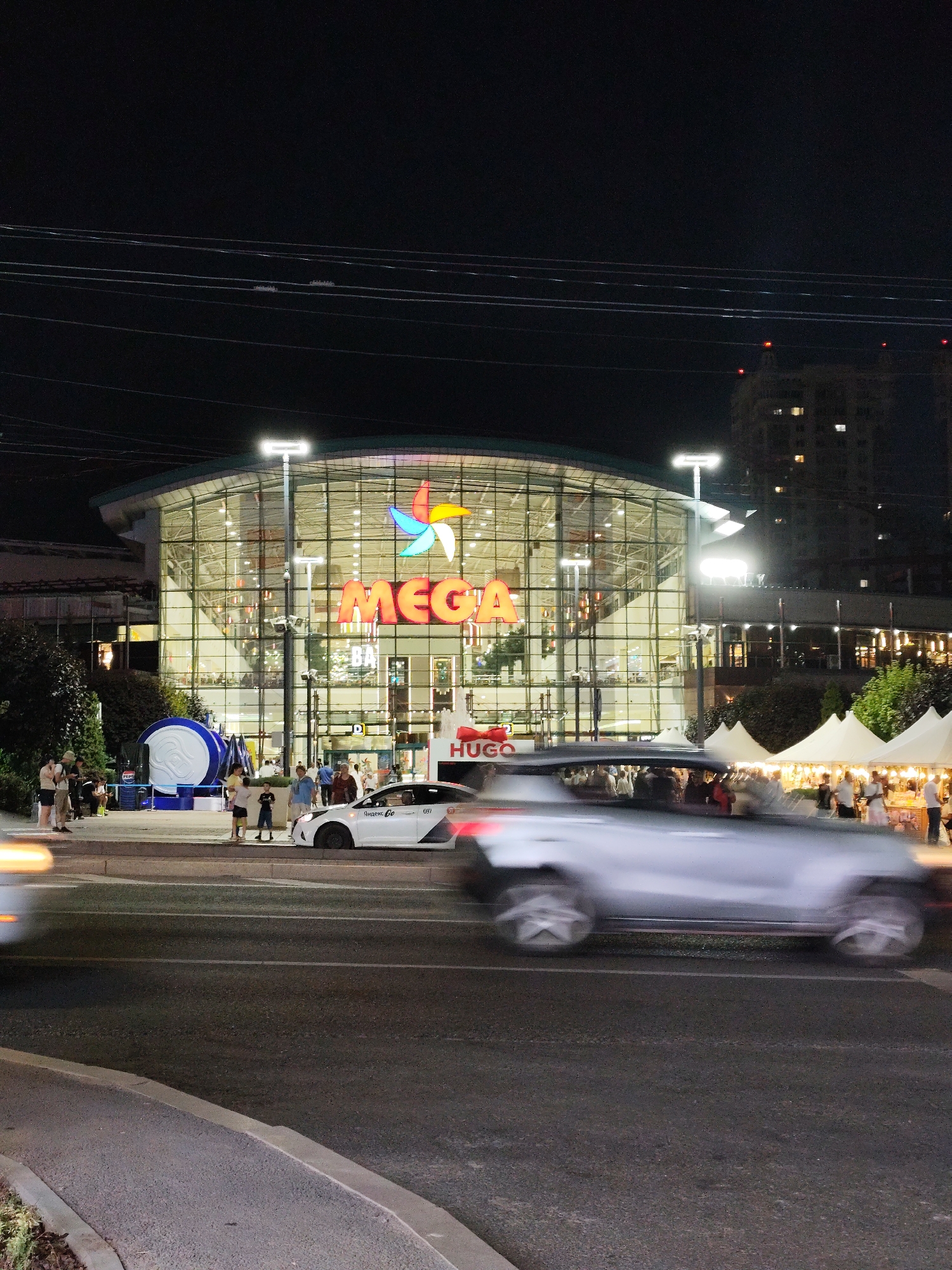 The image shows the "Mega Alma-Ata" shopping and entertainment center at night in Almaty, Kazakhstan. The building features a large glass facade with the illuminated "MEGA" logo and a colorful star logo above it. People are visible near the entrance and on the sidewalk. A white car with "Яндекс Go" branding is parked on the street, and other vehicles are blurred in motion, indicating traffic. To the right of the shopping center, there are white tents set up, possibly for a market or event. The street in front of the building is illuminated by streetlights. The overall scene depicts a busy commercial area at night.