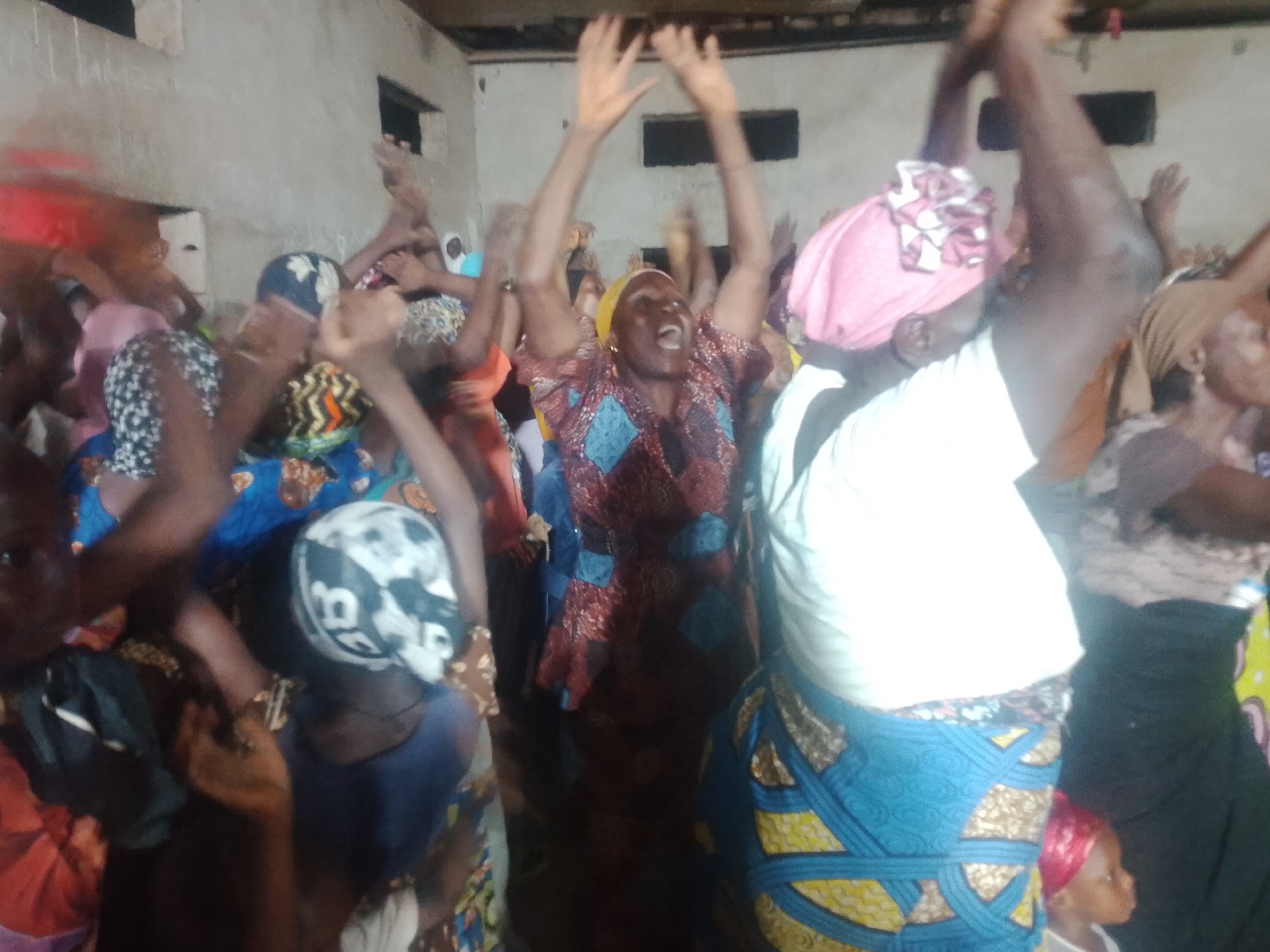 A group of women and at least one child are gathered indoors, with some of them raising their hands in the air. The room appears to be a simple structure with concrete walls and a wooden ceiling. The lighting is dim and appears to be artificial. The individuals are wearing colorful, patterned clothing, including headscarves. The woman in the center of the image is wearing a red and blue patterned dress and has her mouth open, seemingly in a vocal expression. Another woman to her right is wearing a pink headscarf. A child with a red headscarf is visible near the bottom right of the frame. The overall atmosphere appears to be one of excitement or worship.