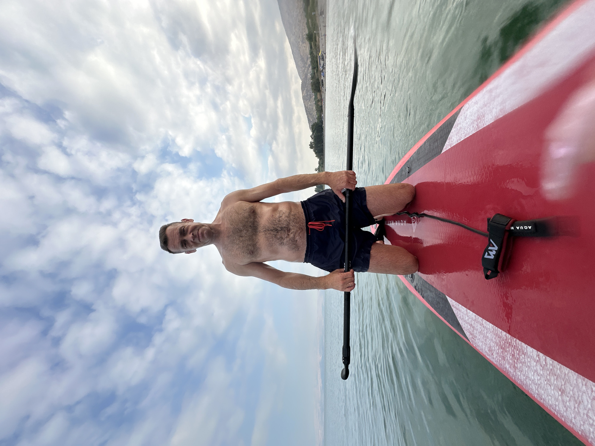 A shirtless man with dark hair and a lean build is kneeling on a red paddleboard in the Sea of Galilee, Emek HaYarden Regional Council, Israel. He is wearing dark blue swim trunks with a red drawstring and holding a paddle across his lap with both hands. The water is calm and appears slightly greenish, with small ripples. The sky is partly cloudy with scattered white and gray clouds. In the background, a distant shoreline with green vegetation and hills is visible, with a few tents and people gathered on the beach. The scene captures a recreational moment on the water under a dynamic sky.