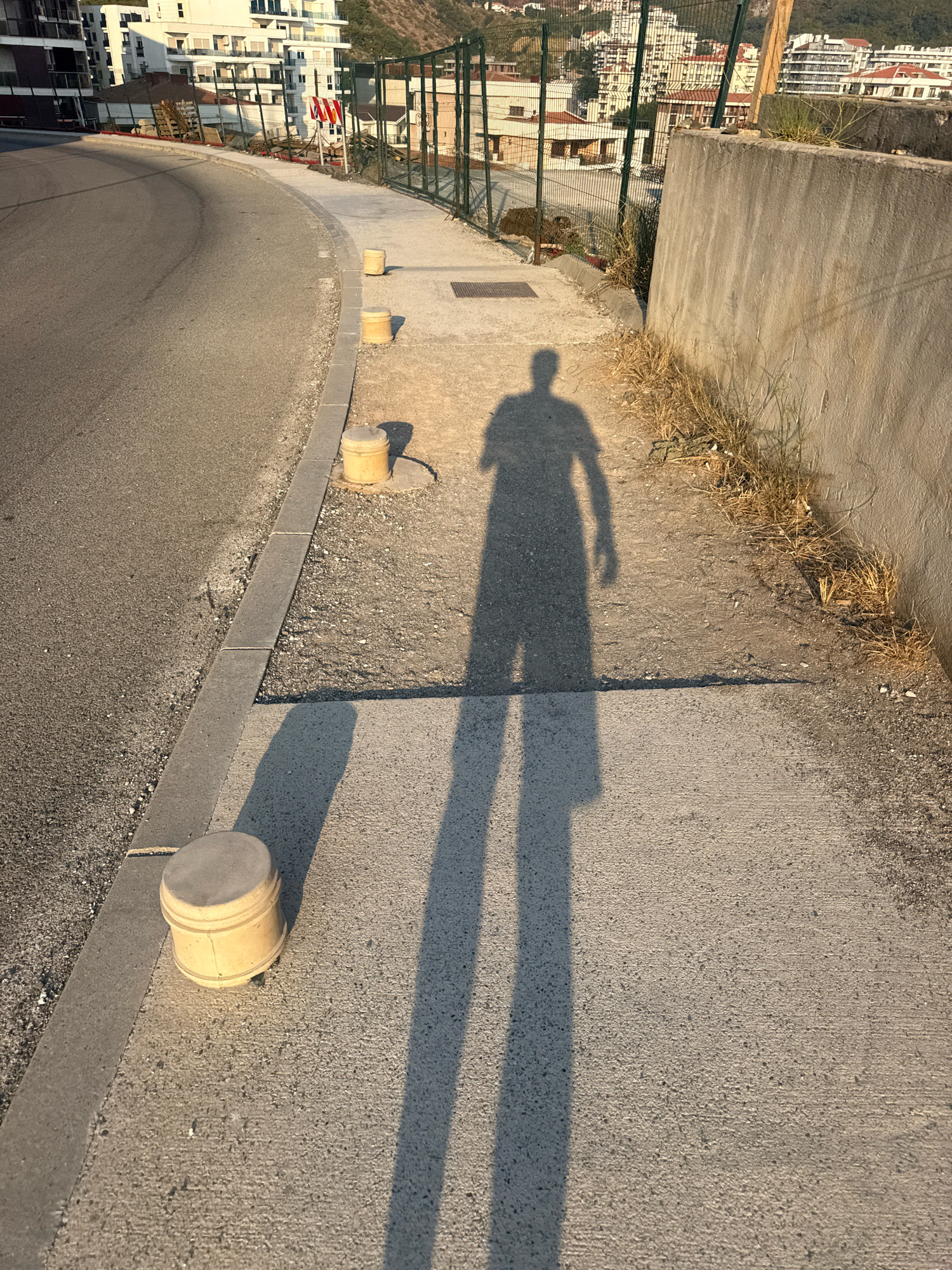 An overhead shot captures a person's shadow cast on a sunlit sidewalk in Boreti, Montenegro. The shadow, elongated by the low-angle sun, extends across the concrete path, with its head and shoulders clearly defined, and its legs stretching out ahead. To the left, a curved asphalt road runs alongside the sidewalk, separated by a concrete curb. Yellow cylindrical bollards are strategically placed along the sidewalk's edge, with one in the foreground and two more further up. In the background, buildings of various heights ascend a hill, some appearing residential and others more commercial. A metal fence runs along the edge of the sidewalk, behind which dry grasses grow. A textured concrete wall forms the right boundary of the sidewalk.