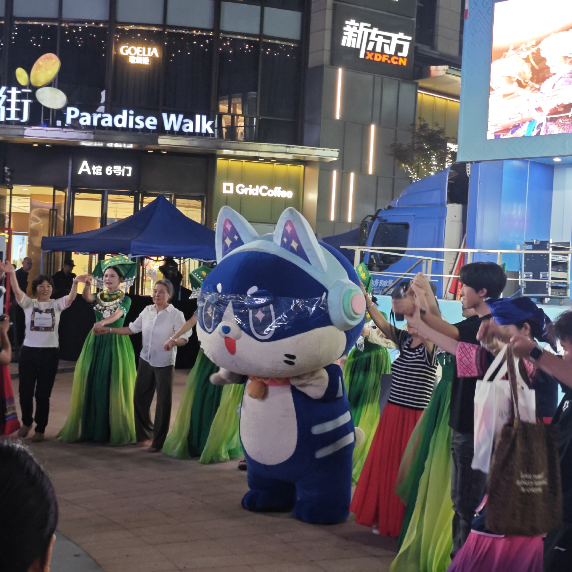 Here is a detailed description of the image:
**Scene:** The image captures a street scene at night in what appears to be a commercial area. A group of people are dancing in a circle, holding hands. The main focal point is a large, blue, cat-like mascot costume in the center.
**People:** A mixed group of individuals are involved in the dance. There is a person wearing traditional green attire with decorative headwear and silver ornamentation. Others are dressed in casual clothes. Several people appear to be holding up their hands, possibly in a dance or celebratory gesture.
**Objects:**
* Buildings with visible signs like "Paradise Walk", "GOELIA", "XDF.CN" (translated from Chinese), and "Grid Coffee" are visible.
* A stage-like structure can be seen in the background, and a blue truck is parked nearby.
* Various shopping bags are held by some individuals.
* The mascot costume is a large, blue figure with cat-like features, including ears, and wearing what looks like headphones.
**Actions/Interactions:** The individuals are engaged in a group dance, holding hands in a circle. There seems to be an interaction between the crowd and the mascot.
**Environment:** The setting is an outdoor urban area, likely in the evening or at night, given the lighting.
**Location:** The image includes signs with English and Chinese text, suggesting the location is in China. Given the information in the signs in the picture, it is likely that this photo was taken in Yuzhong District in China at Paradise Walk shopping mall.