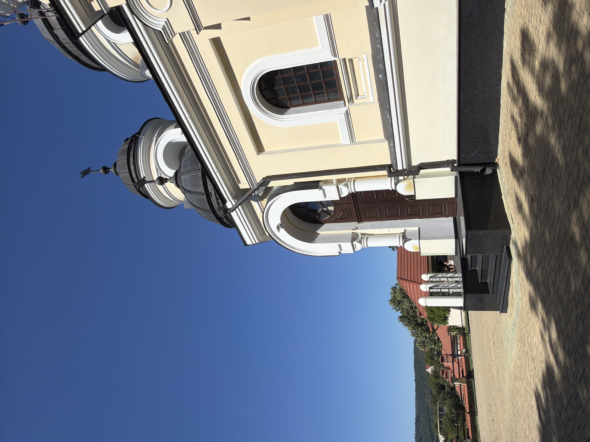 A medium shot captures the exterior of a yellow-painted building, likely a church, against a clear blue sky in Căpriana, Moldova. The building features dark domed roofs topped with crosses and arched windows with dark panes. A dark brown door is visible under a white arched entranceway. To the left, red-tiled roofs of other buildings and lush green trees are visible in the background, with a sloping, tree-covered hill in the far distance. The foreground is paved with tan brickwork, casting striped shadows from an unseen source. A black granite staircase leads up to the entrance, with a black drainpipe running down the side of the building.